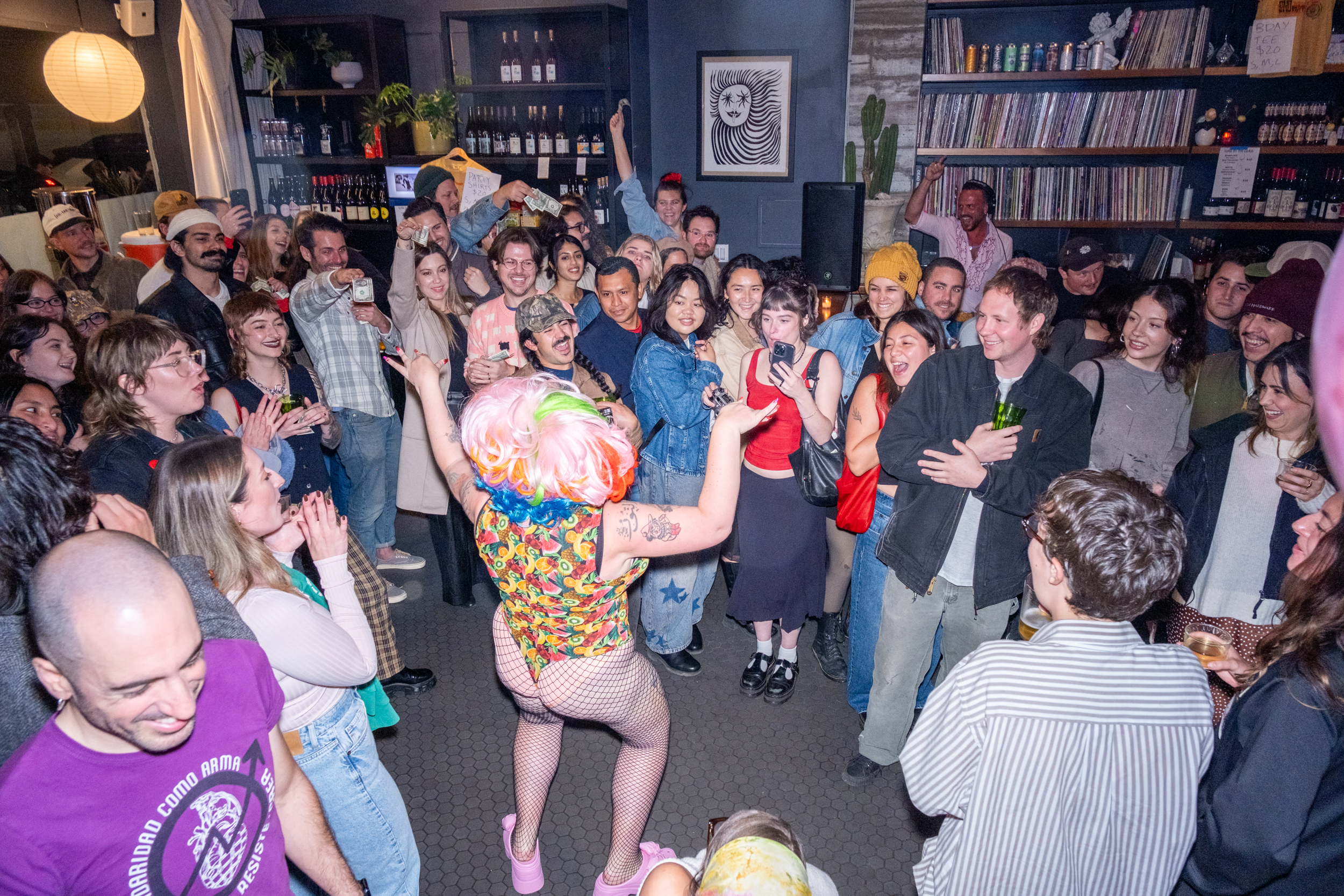 A lively crowd smiles and claps around a performer with colorful, curly hair and vibrant outfit, in a cozy room with shelves of records and bottles.