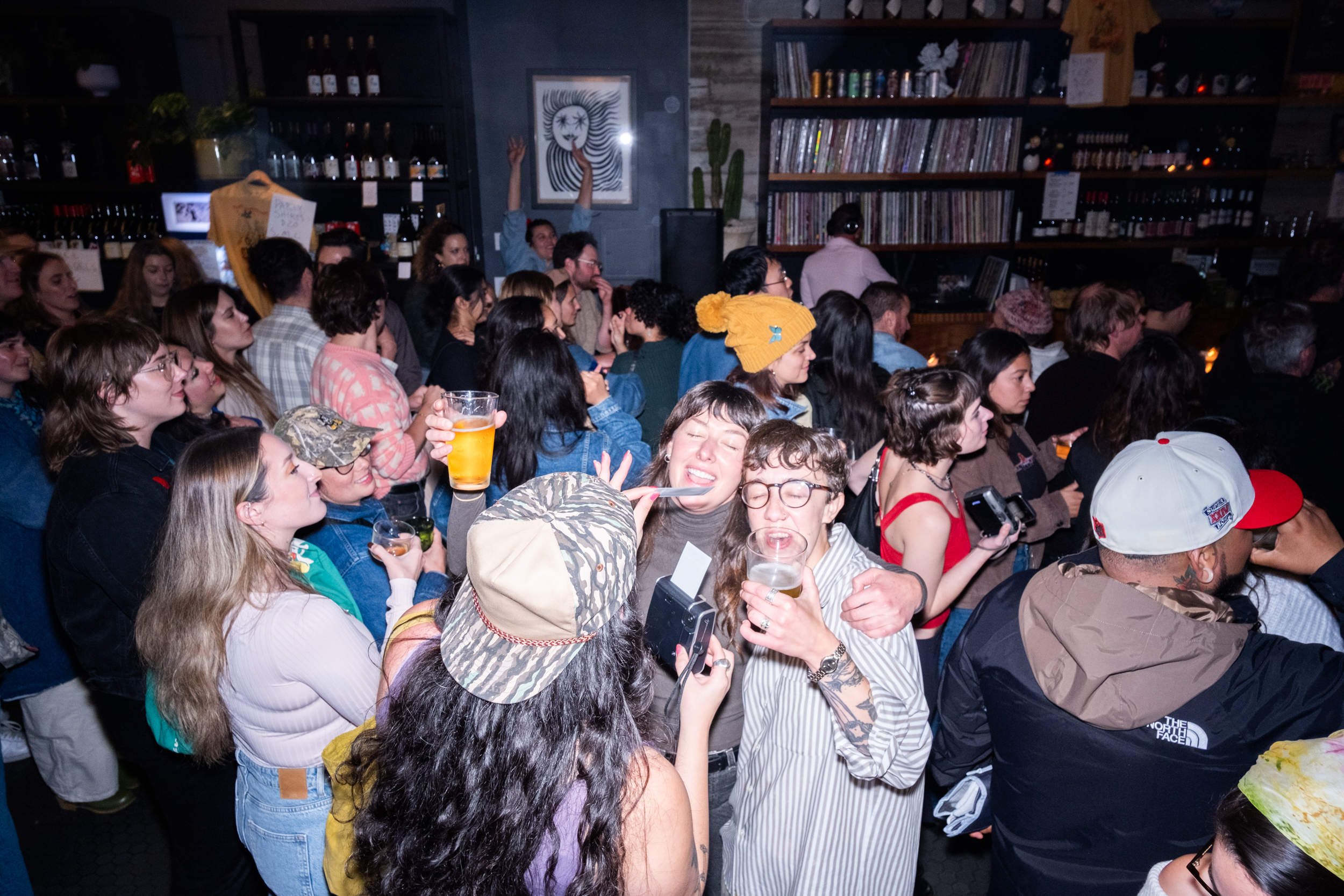 A lively bar scene with people laughing, holding drinks, and capturing moments. Shelves filled with records and bottles are seen in the background.