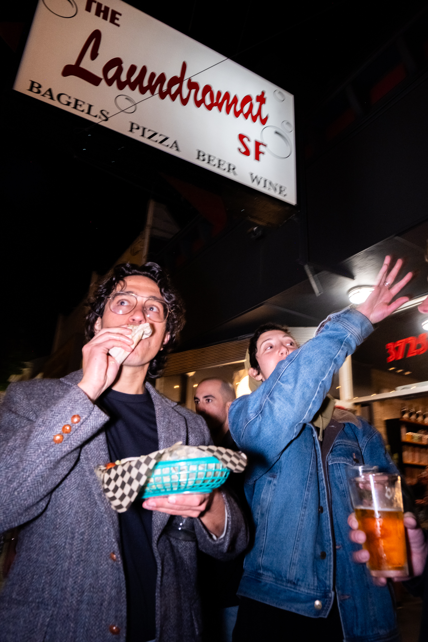 Two people stand at night near a sign for &quot;The Laundromat SF,&quot; eating and drinking; one is holding a food basket, the other a beer. Another person is in the background.