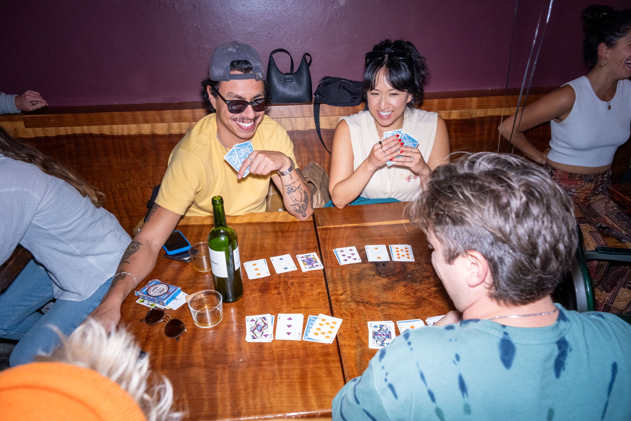 People are playing cards at a wooden table, laughing and enjoying themselves. There's a bottle and some drinks on the table, and the atmosphere is relaxed and fun.