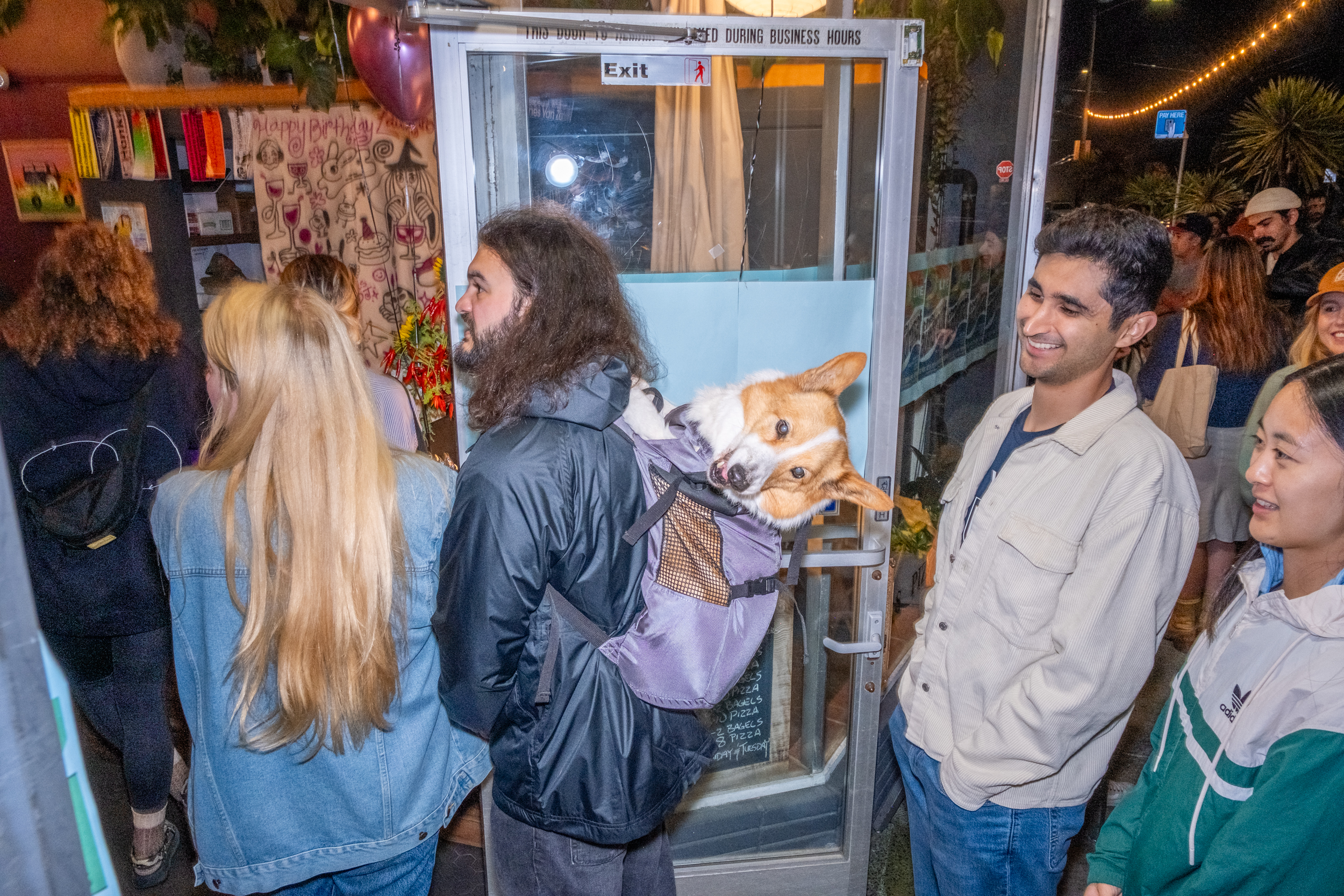 A crowd is gathered at a doorway. A man with long hair carries a corgi in a backpack, drawing smiles from those around him. Balloons and a birthday banner are visible.