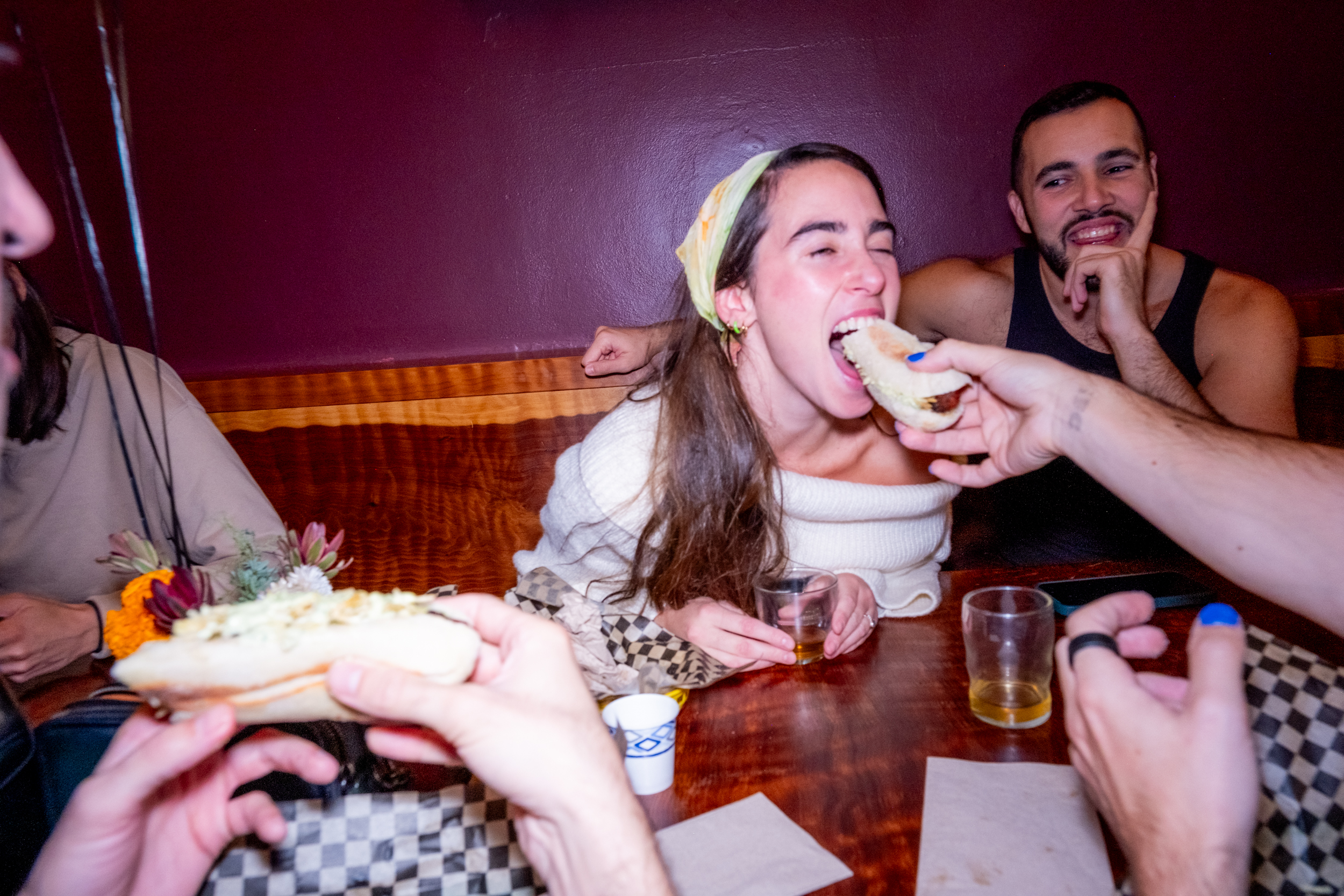 A woman is humorously trying to eat a hot dog being held by another person, while several people sit around them in a lively, playful atmosphere.