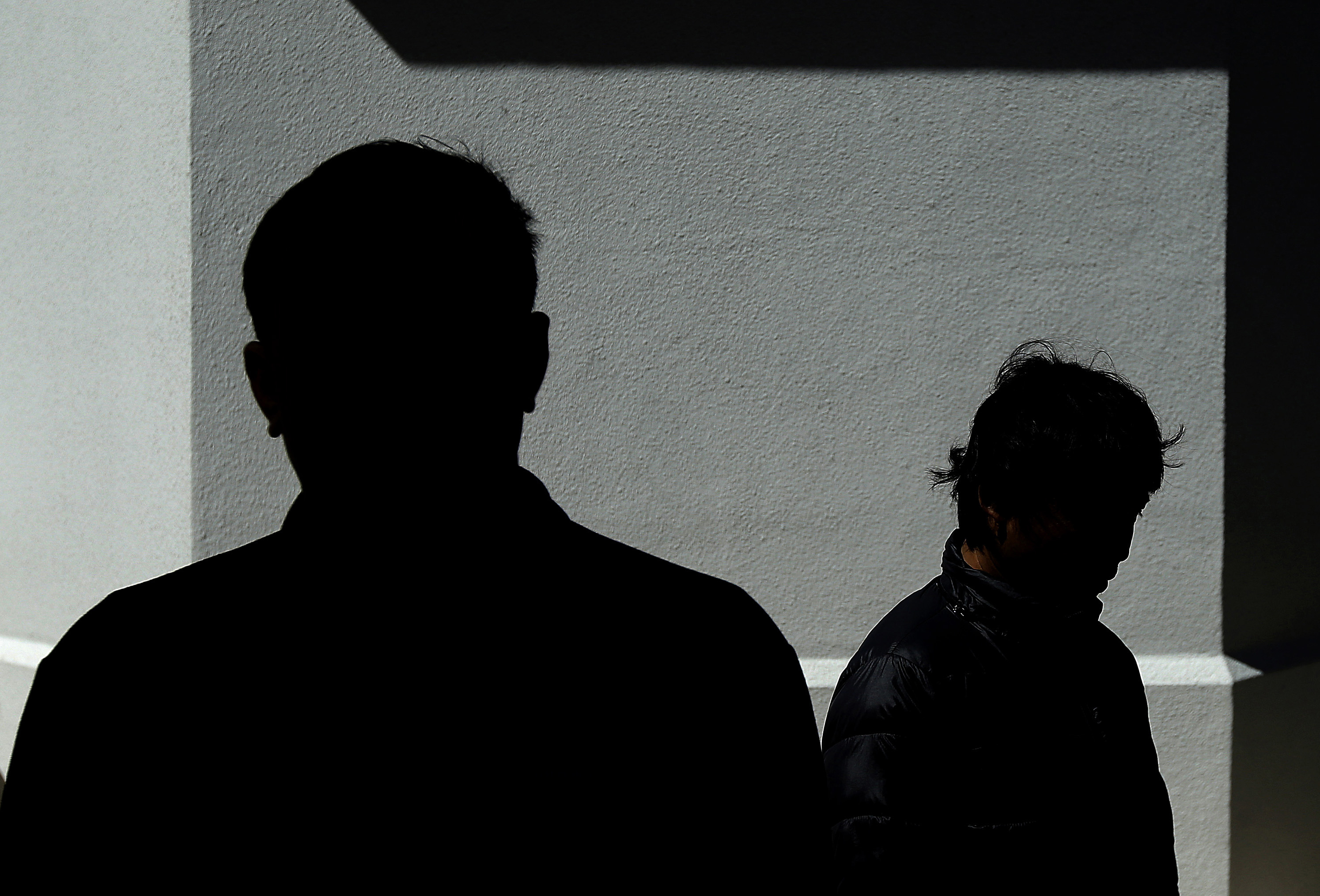 Two silhouetted figures stand against a light gray wall in shadow, creating a dramatic contrast with the bright background.