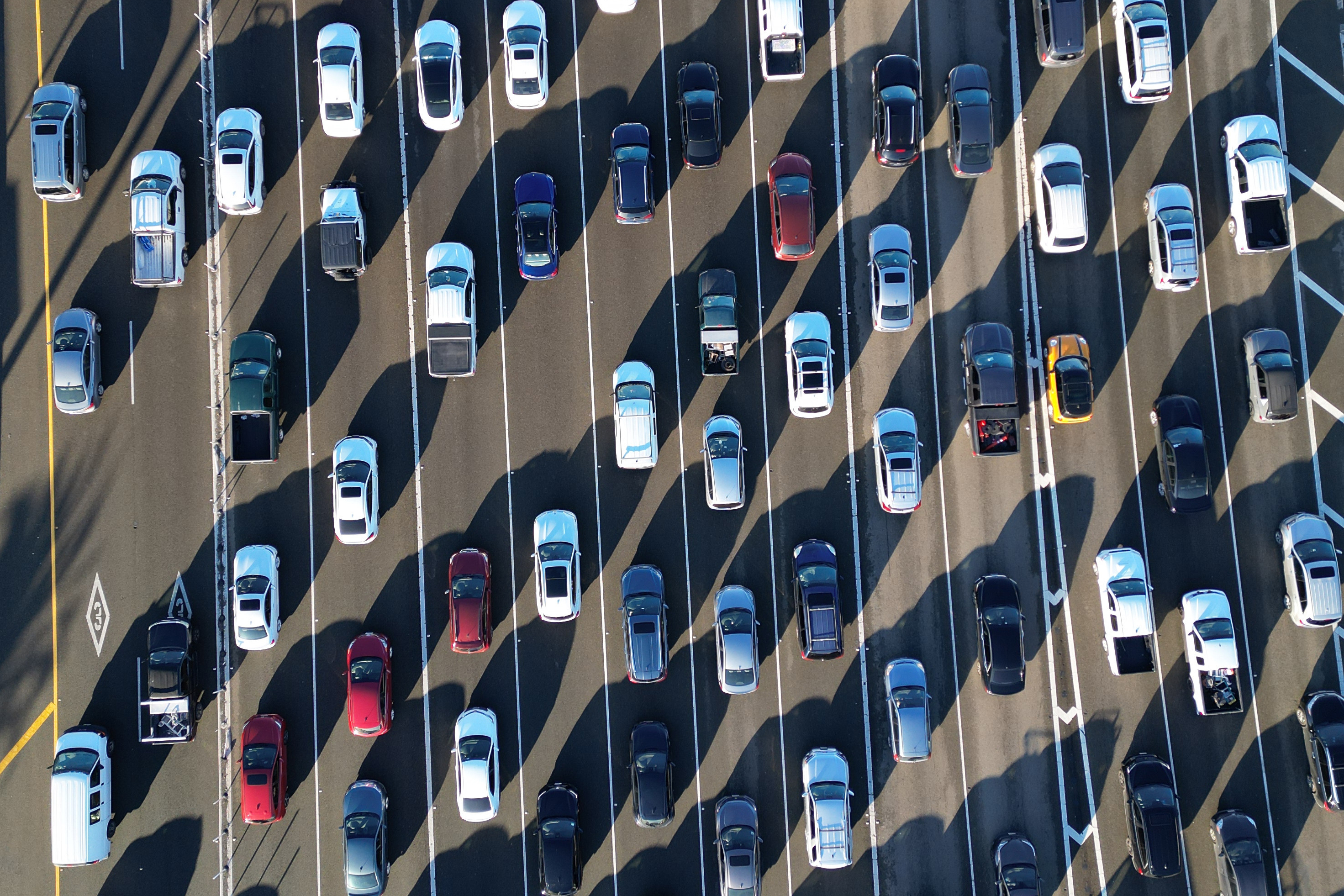 The image shows multiple lanes of traffic from above, with rows of cars and trucks in various colors, creating a pattern of parallel lines and shadows.