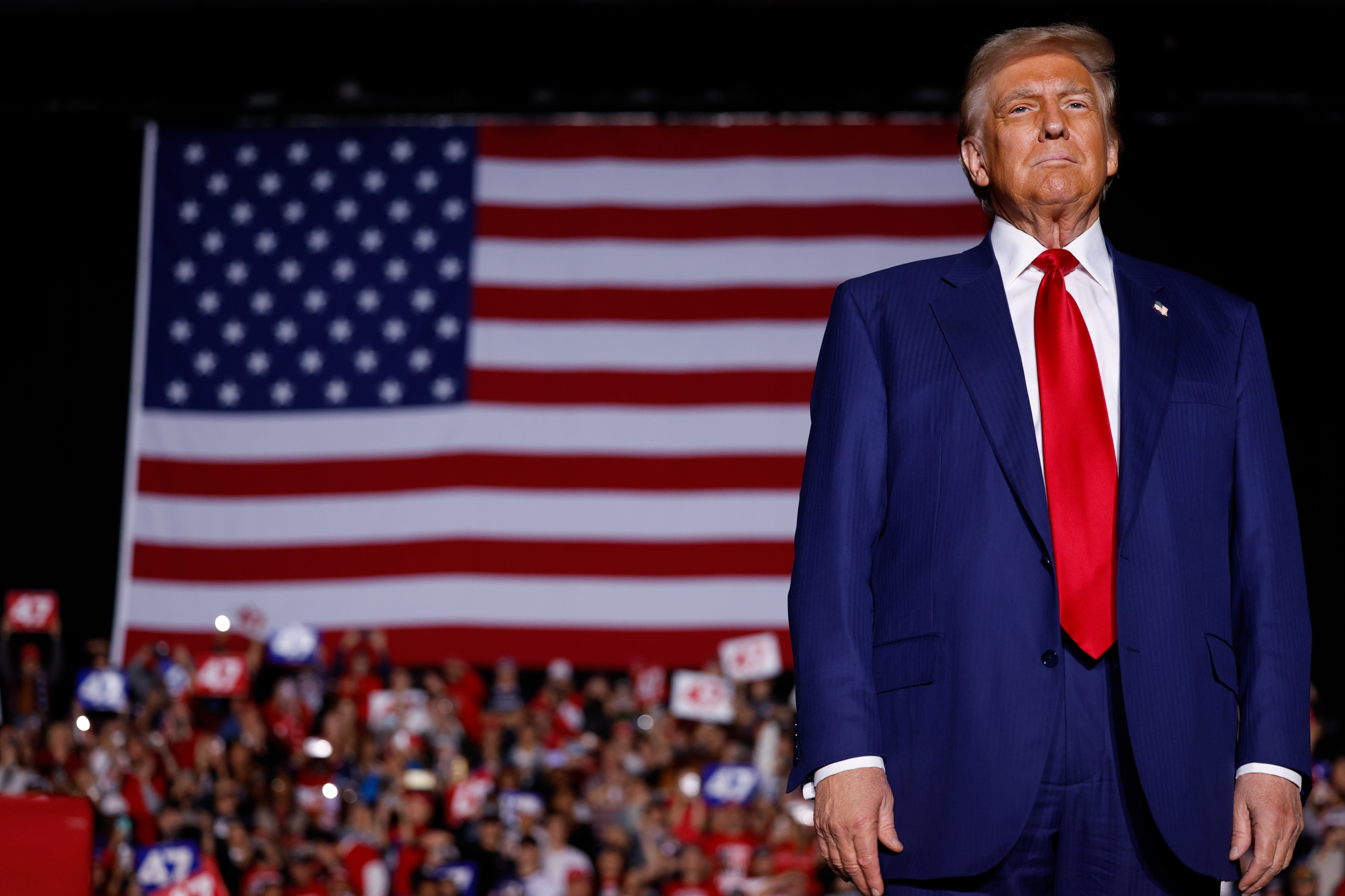 A man in a blue suit and red tie stands confidently in front of a large American flag, with a cheering crowd holding signs in the background.