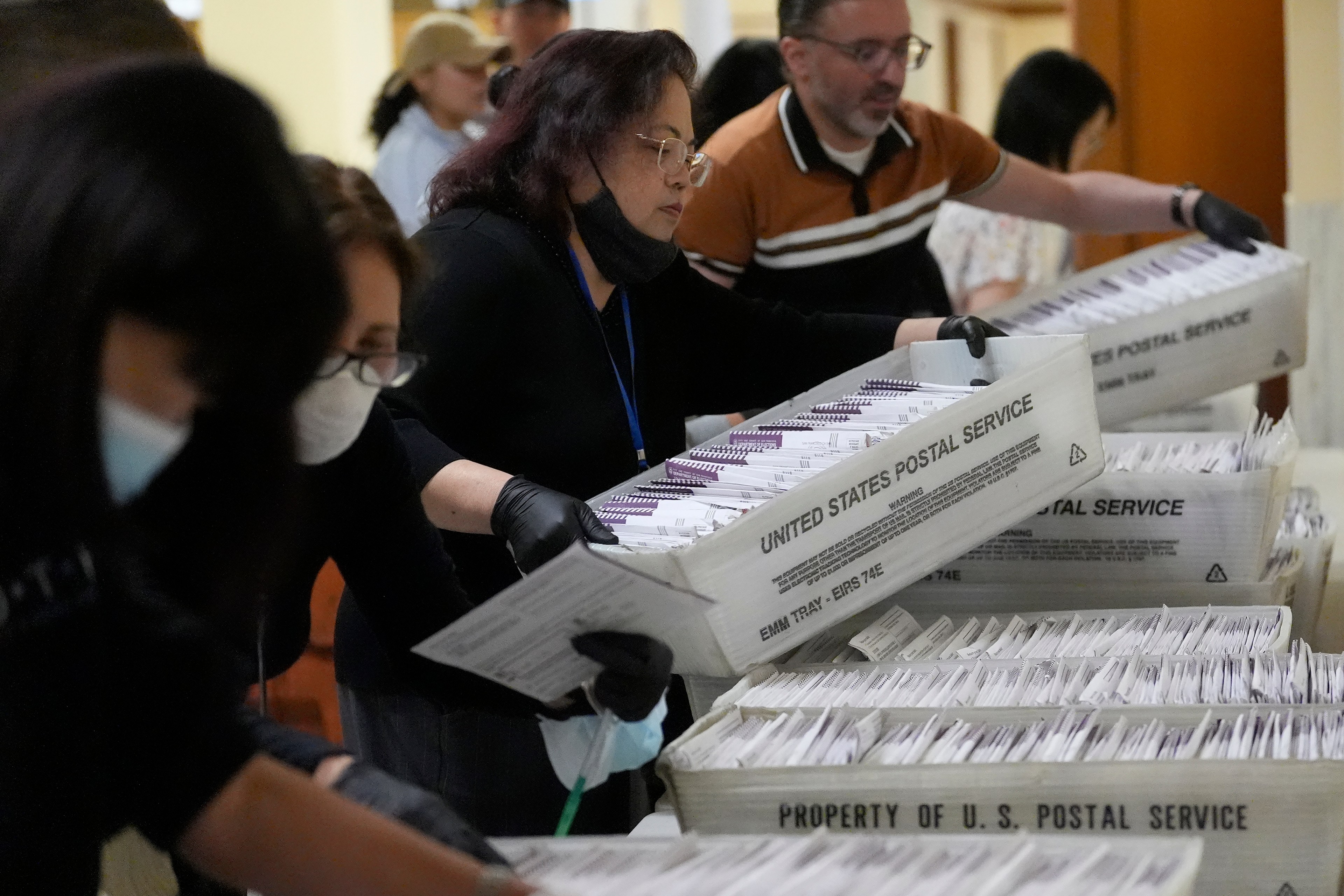 People are sorting and handling stacks of envelopes in USPS trays, likely mail ballots. They wear masks and gloves, working closely together.