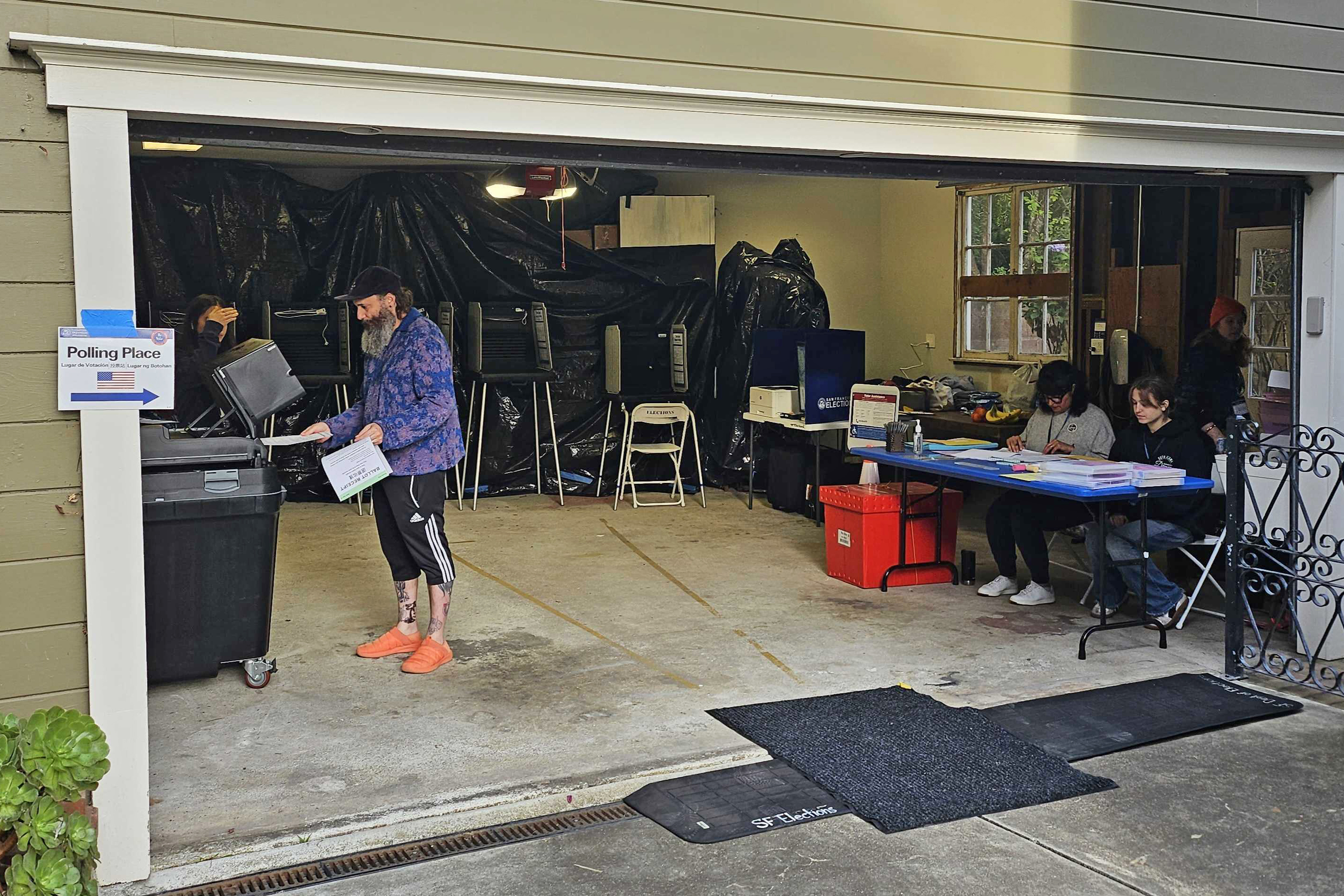 A man is voting in a garage turned into a polling place with a sign outside. Inside, two women sit at a table handling paperwork, while voting booths line the back wall.