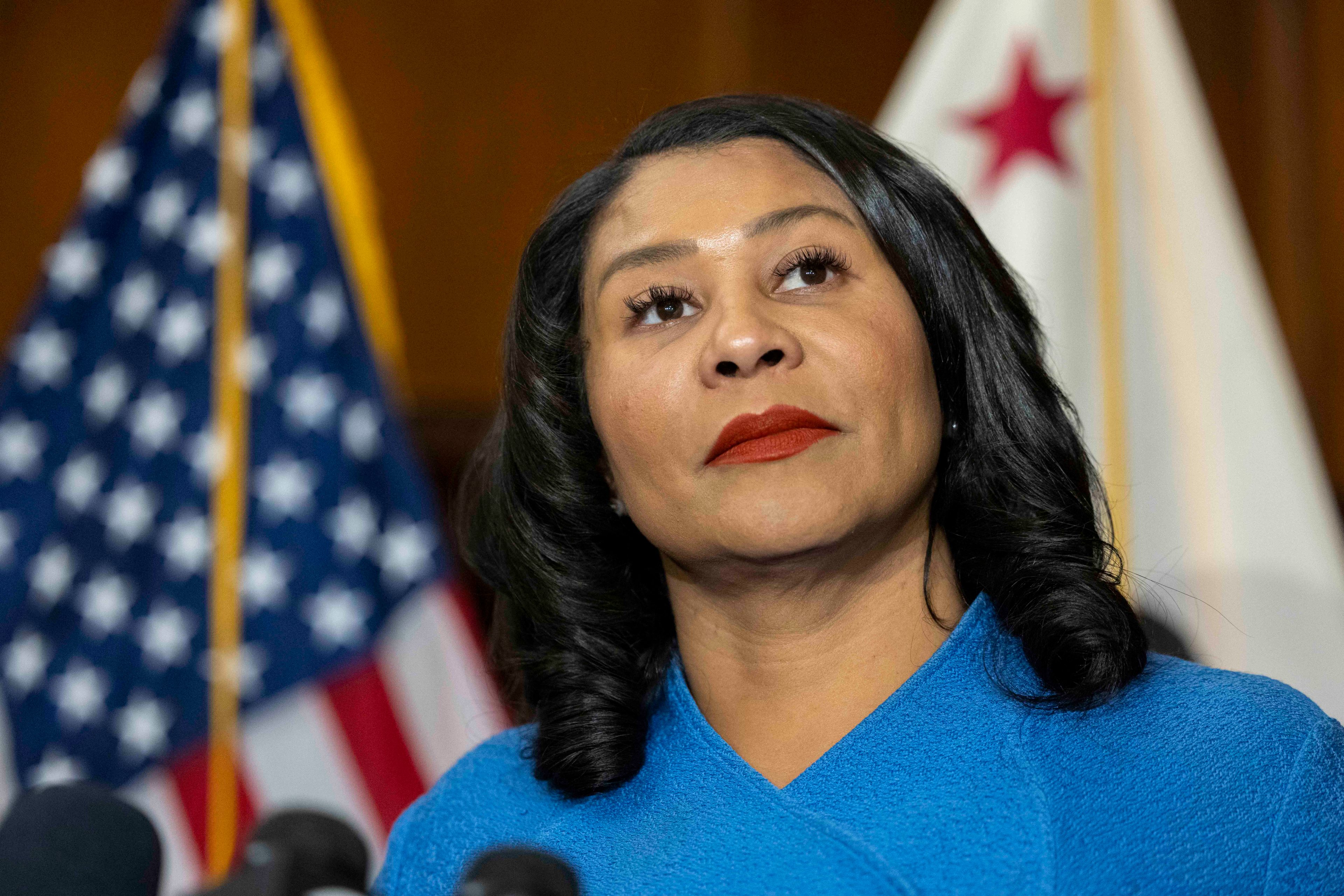 A person in a blue outfit stands before microphones, with U.S. and California flags behind, appearing thoughtful and composed.