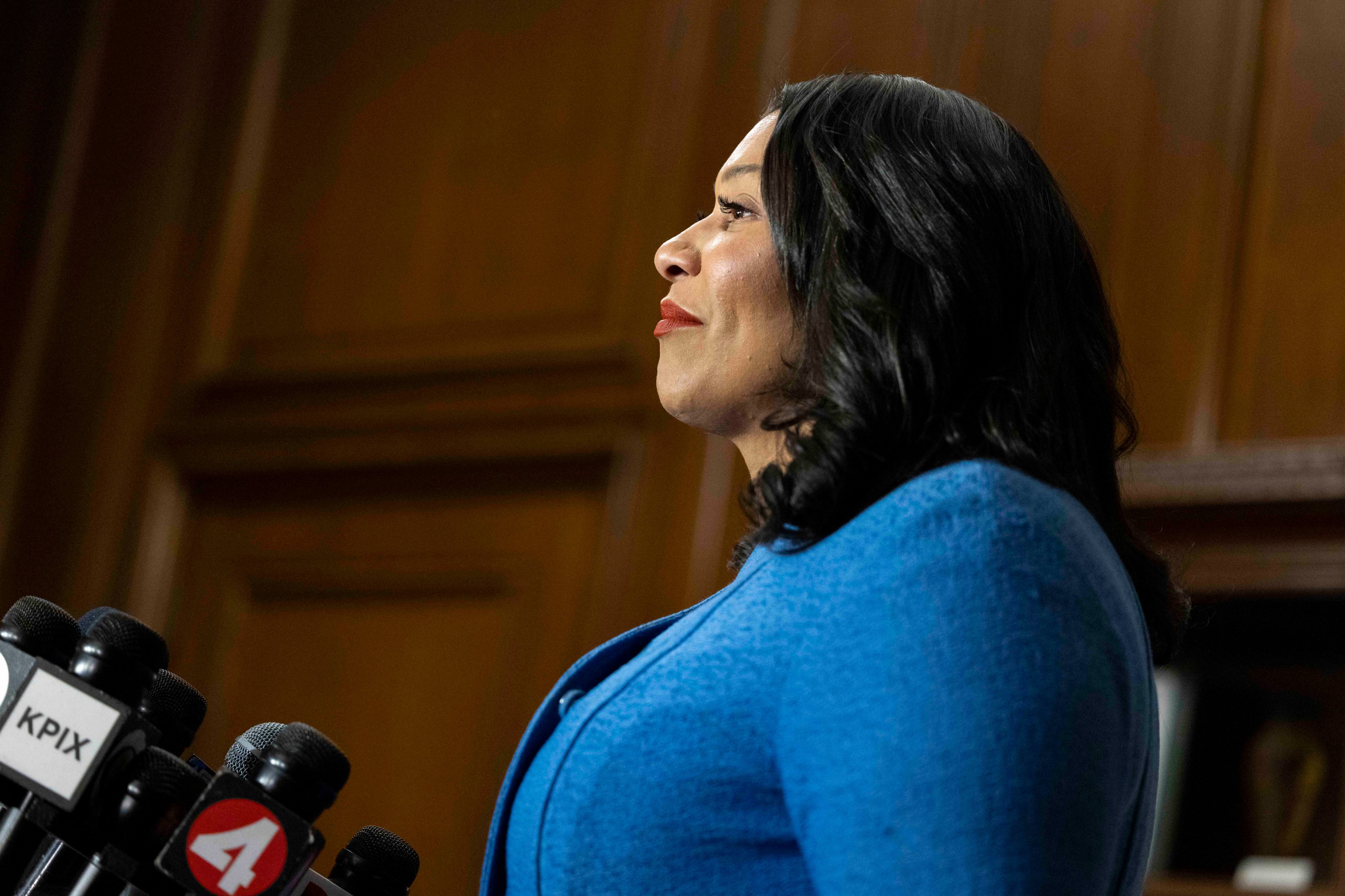 A woman in a blue outfit stands at a podium with multiple microphones. She appears to be speaking or addressing an audience in a formal setting.