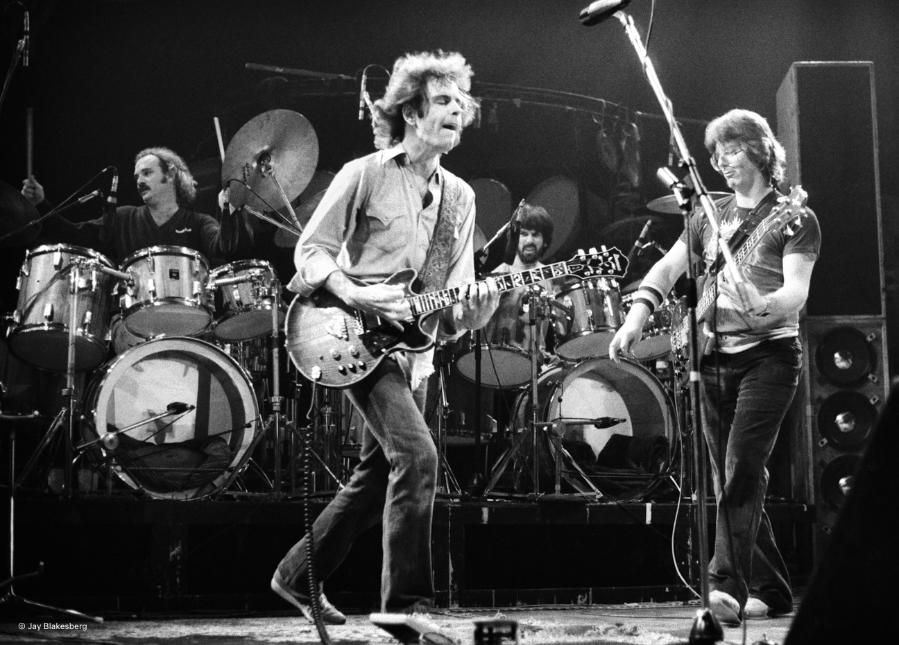 A black-and-white image shows three musicians performing on stage. Two are playing guitars passionately, while a drummer plays energetically in the background.