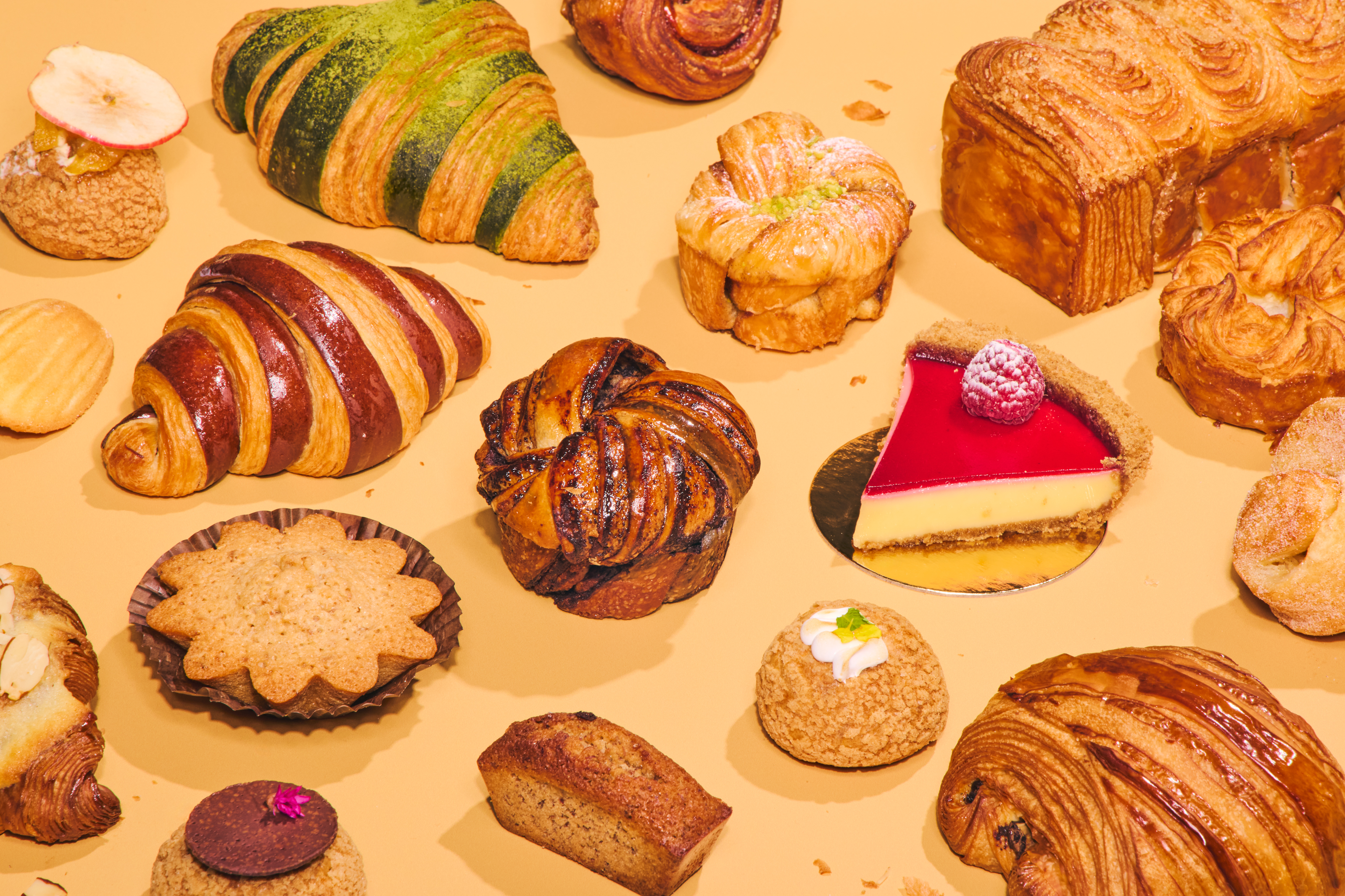 An assortment of pastries, including croissants, tarts, and cakes, are displayed on a beige surface. A slice of raspberry cheesecake is prominently featured.