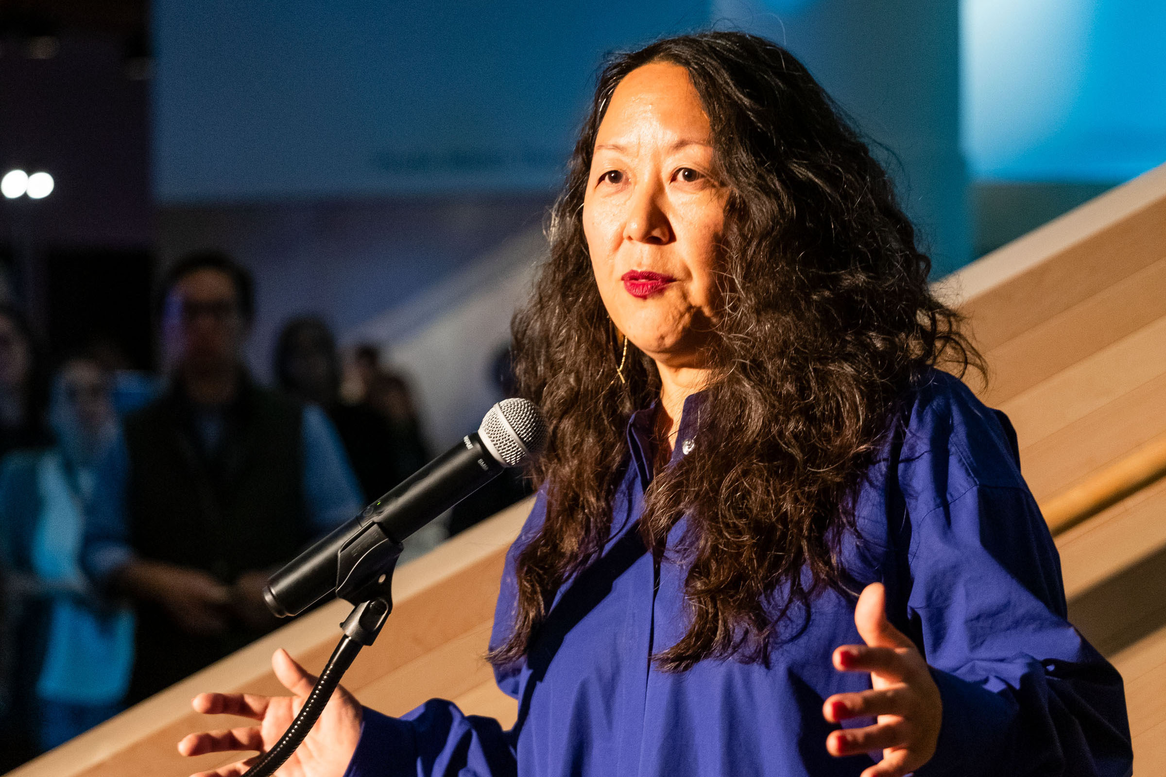 A woman with long wavy hair speaks into a microphone. She's wearing a blue shirt, gesturing with her hands, against a blurred background of people and soft lighting.