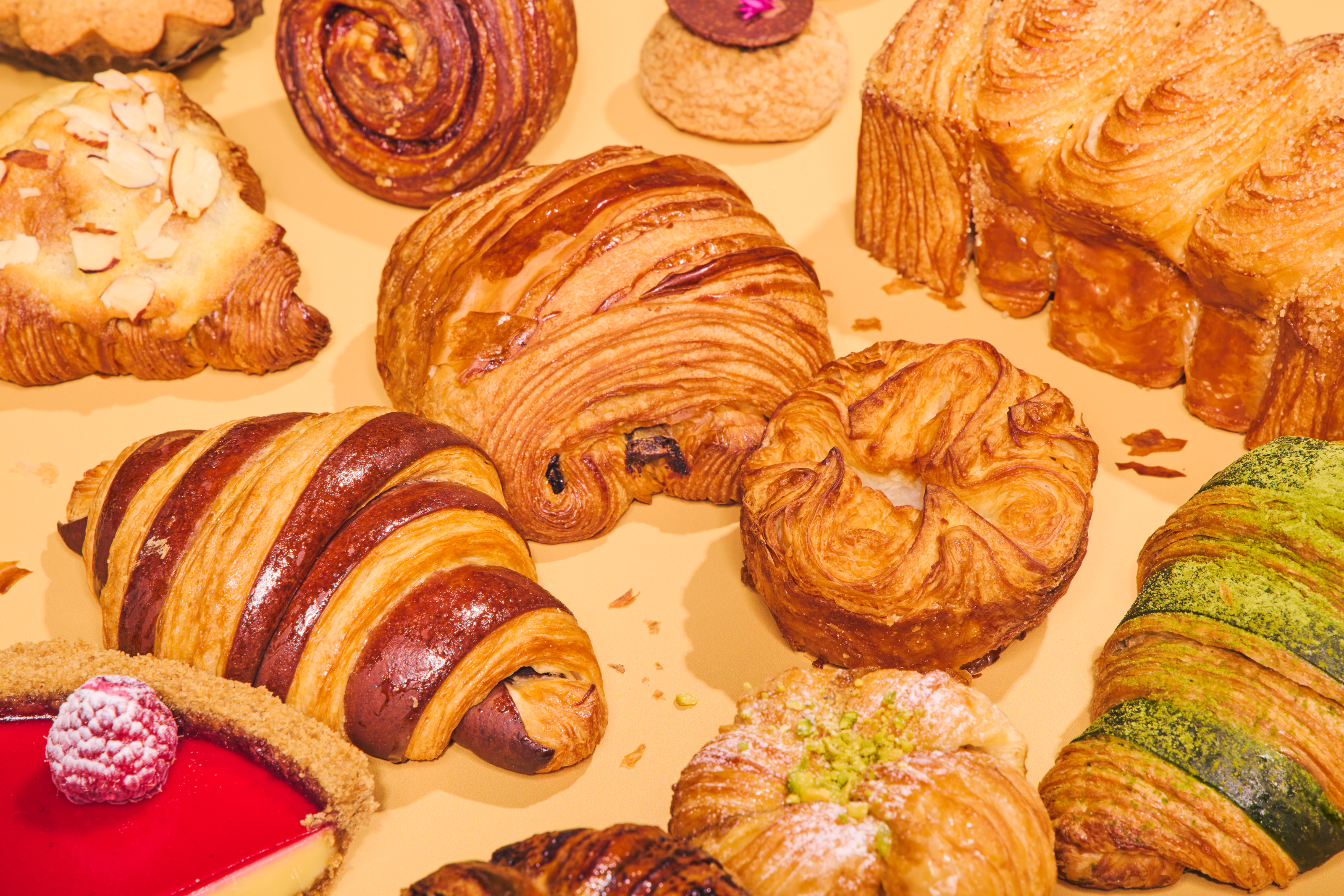 An assortment of pastries on a beige background, including croissants, a swirl roll, a pistachio-topped pastry, and a raspberry dessert, showcasing flaky textures.