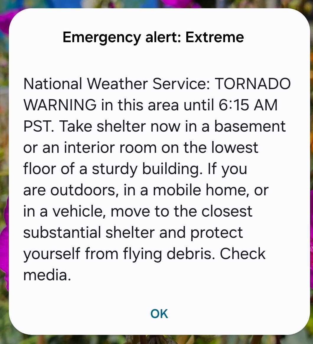 A smartphone screen displays a tornado warning alert, advising immediate shelter due to extreme weather. The background shows colorful flowers.