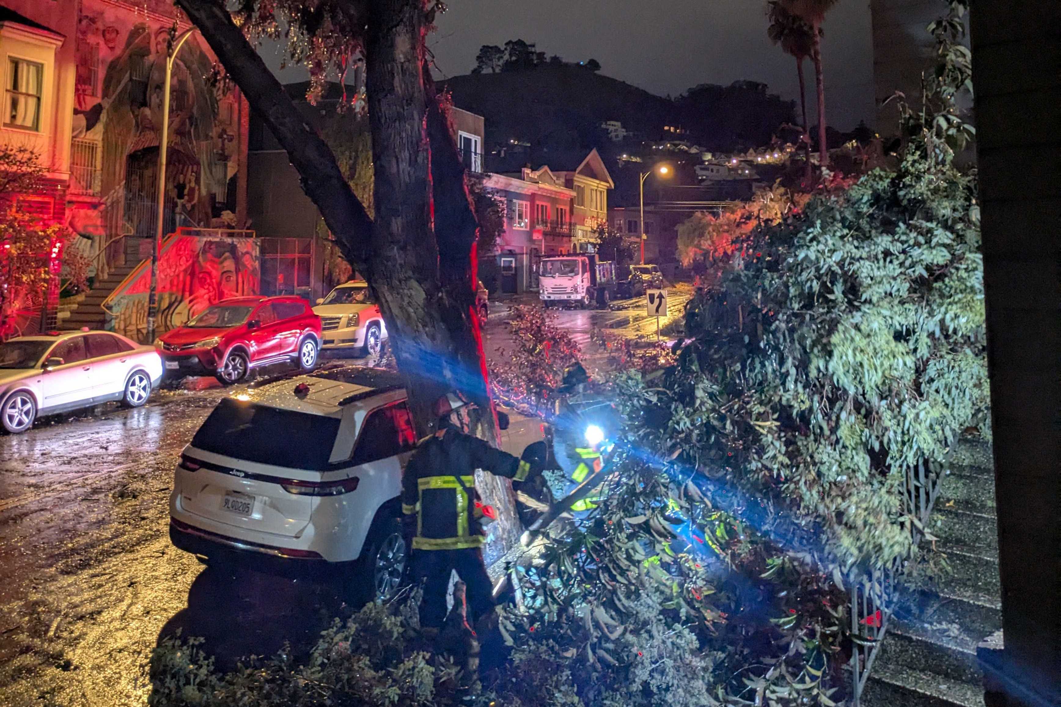 A large tree has fallen onto a white SUV on a dark, rainy street. Emergency workers in reflective gear are clearing the debris. Streetlights illuminate the scene.