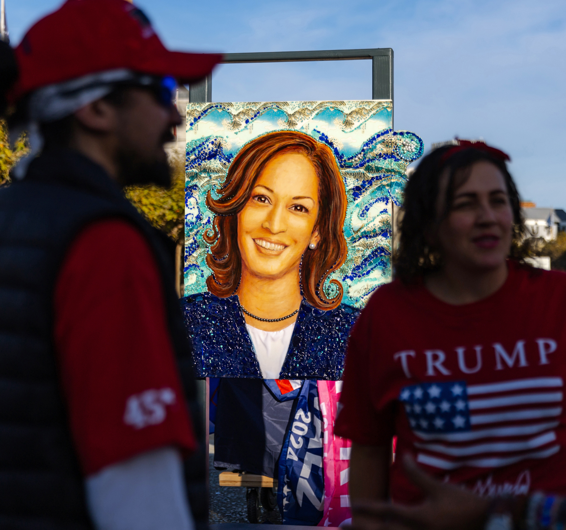 The image shows a vibrant painting of a smiling woman against a wave-patterned background. Two people in red pro-Trump attire stand in front of the artwork.