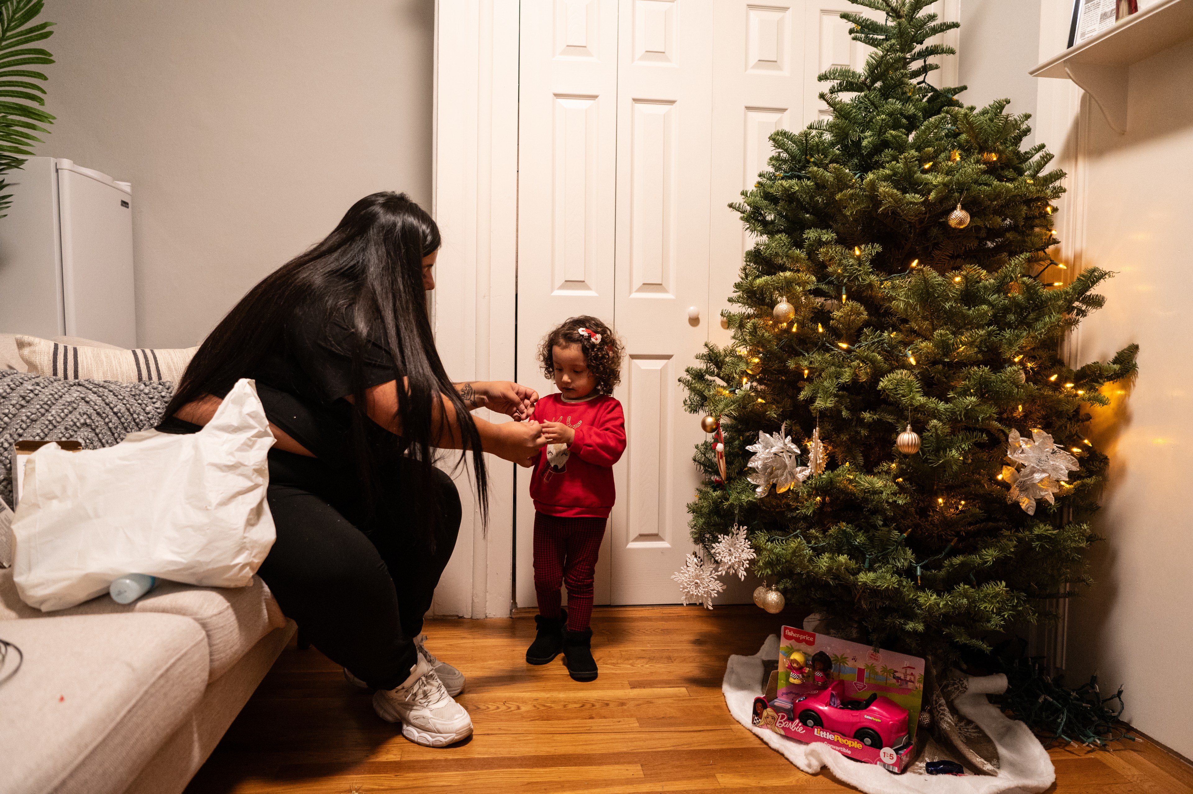 A woman and child decorate a lit Christmas tree with ornaments. A toy car sits under the tree. They're beside a couch with a white bag on it.