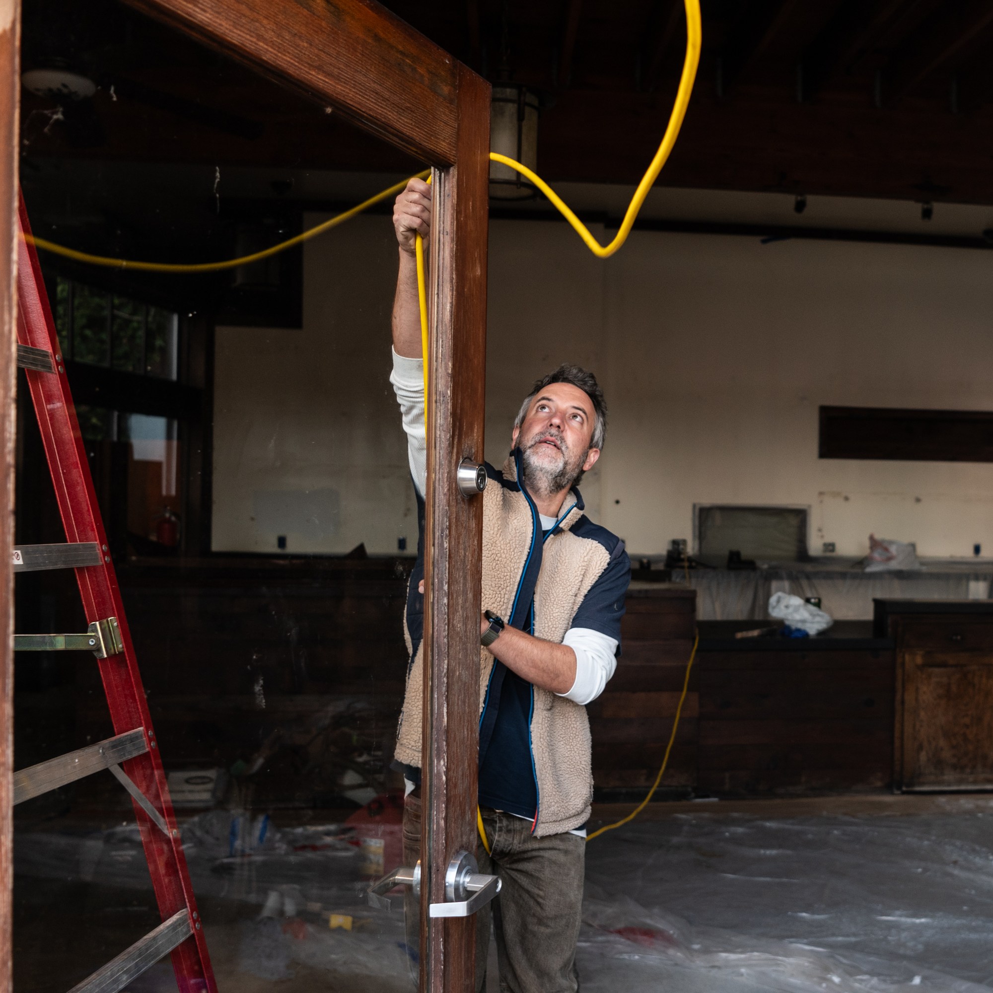 A man is inspecting a wooden door frame inside a room under renovation, holding a yellow cable. A red ladder and plastic coverings are visible in the background.