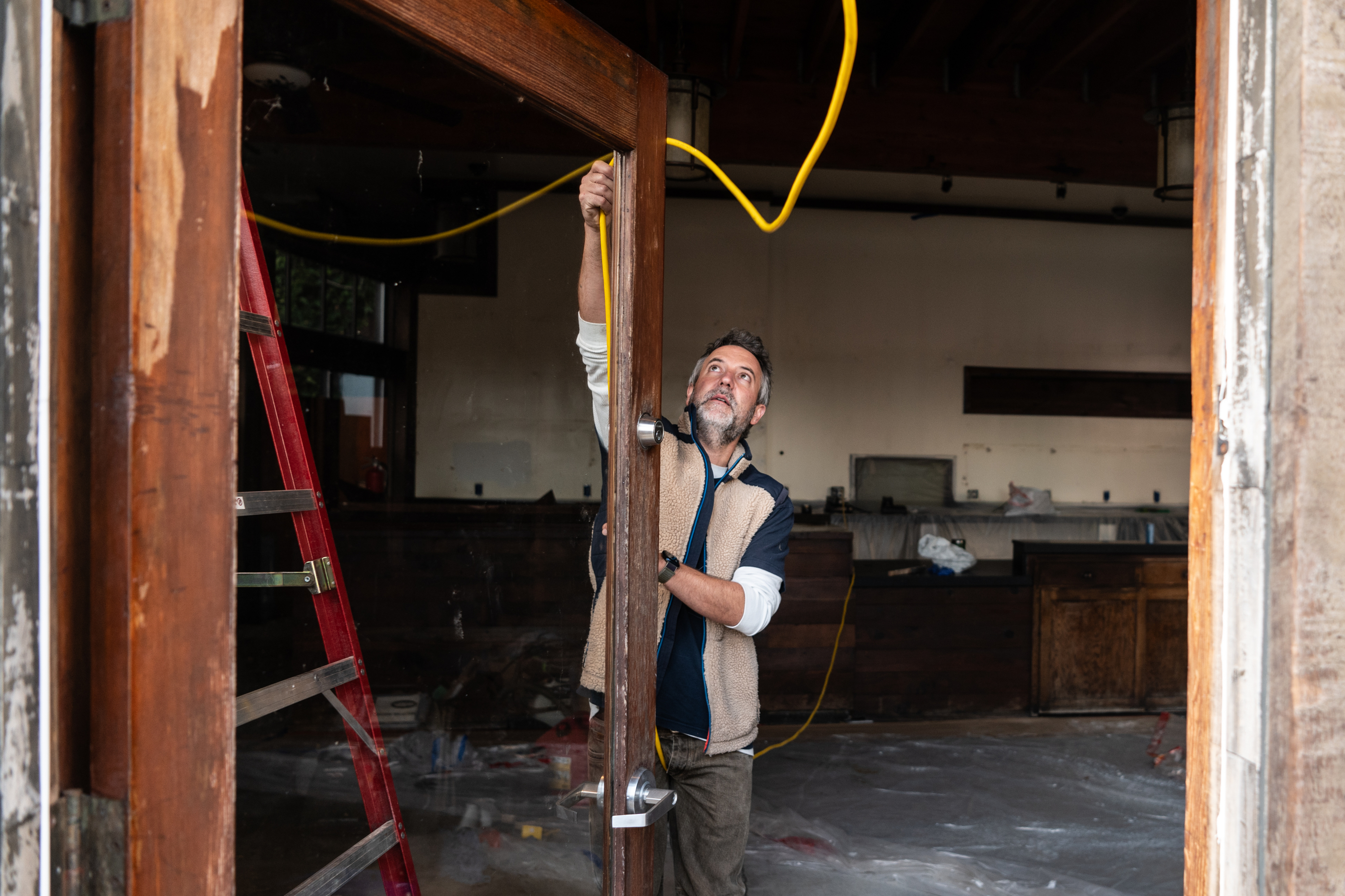 A man is inspecting a wooden door frame inside a room under renovation, holding a yellow cable. A red ladder and plastic coverings are visible in the background.