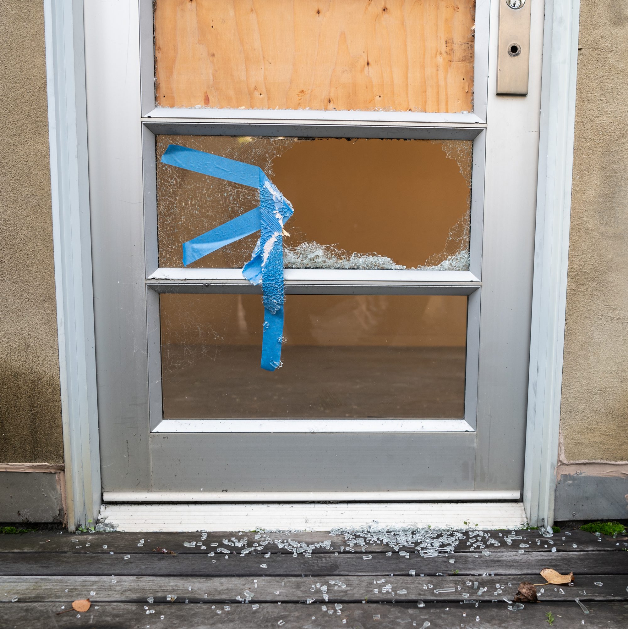 A door with a broken glass panel is patched with blue tape. Shattered glass is scattered on the wooden floor below.