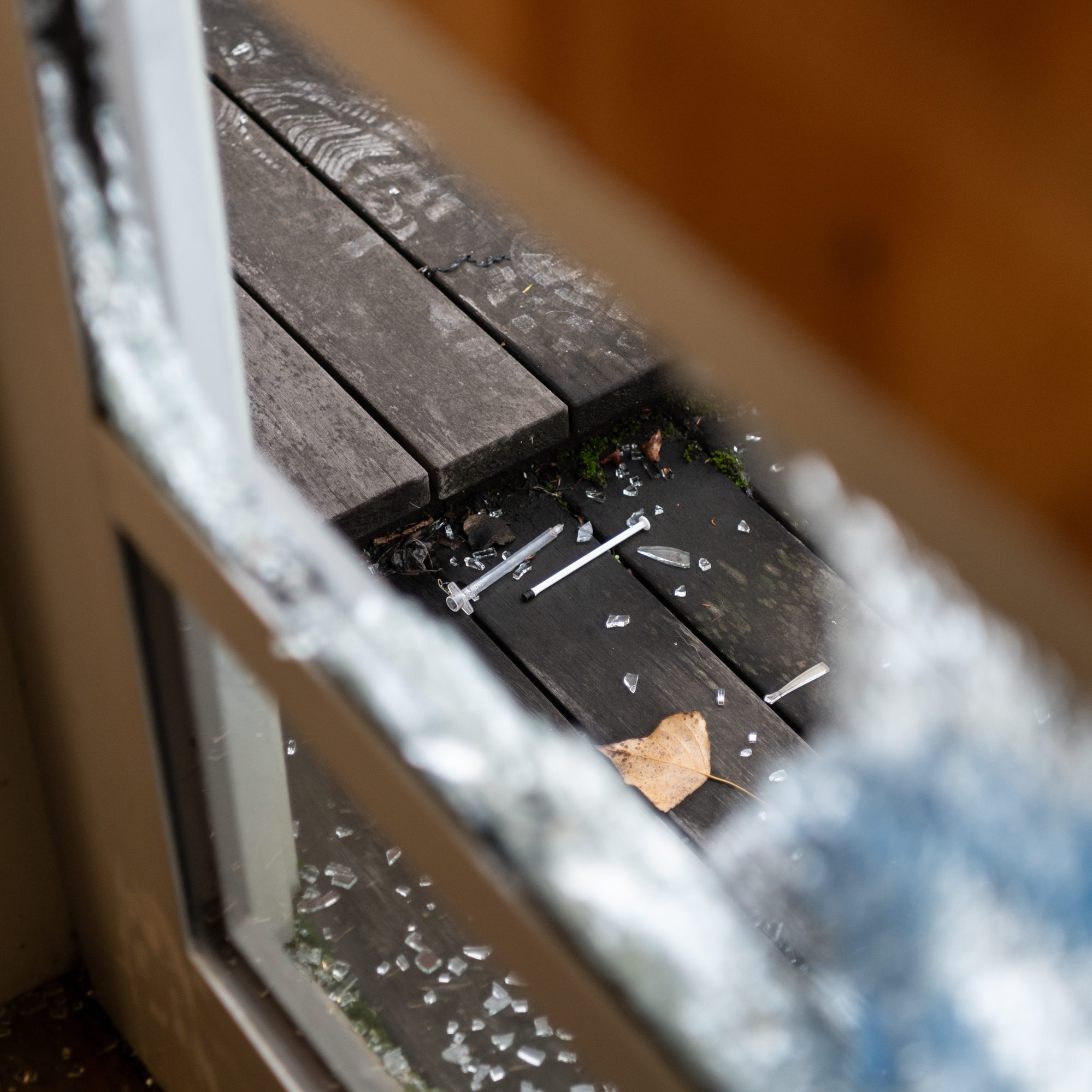 A broken window with shattered glass surrounding it. Below, a wooden floor outside has more glass shards, a needle, a few nails, and a dry leaf.