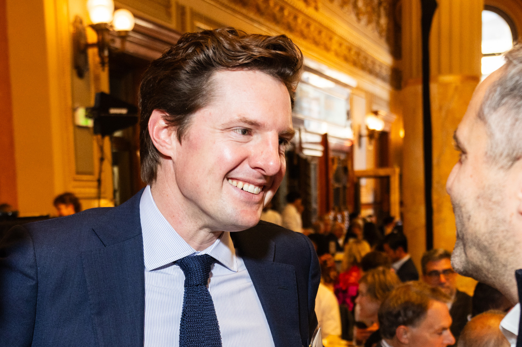 A man in a suit is smiling and conversing with another person in a room filled with people, featuring ornate lighting and architectural details.