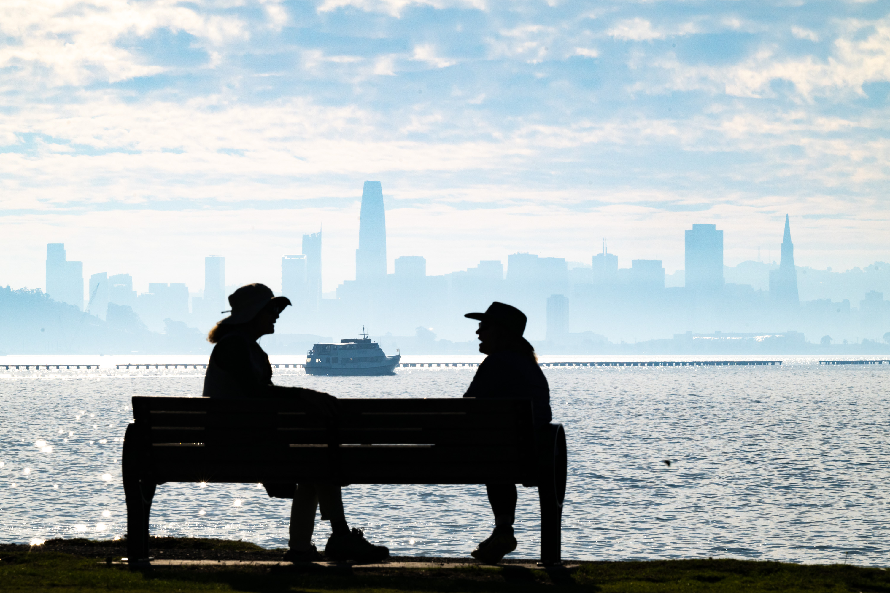 Two people in hats sit on a bench with a misty city skyline and a ferry on the water in the background.