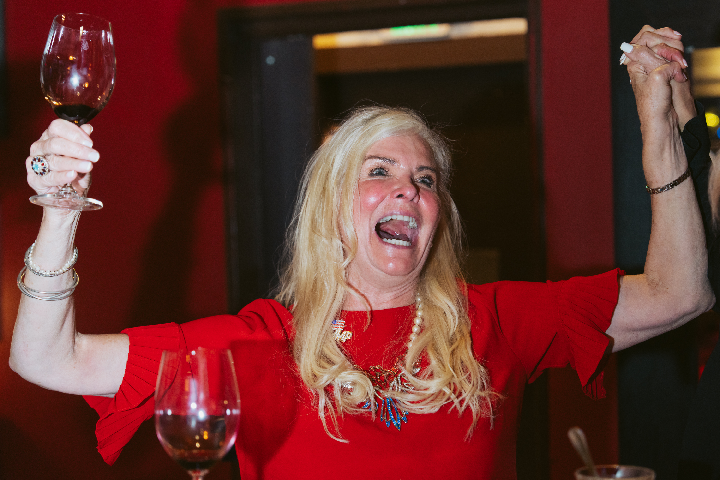 A joyful woman in a red dress holds up a glass of wine and someone's hand, smiling exuberantly, in a warmly lit room.