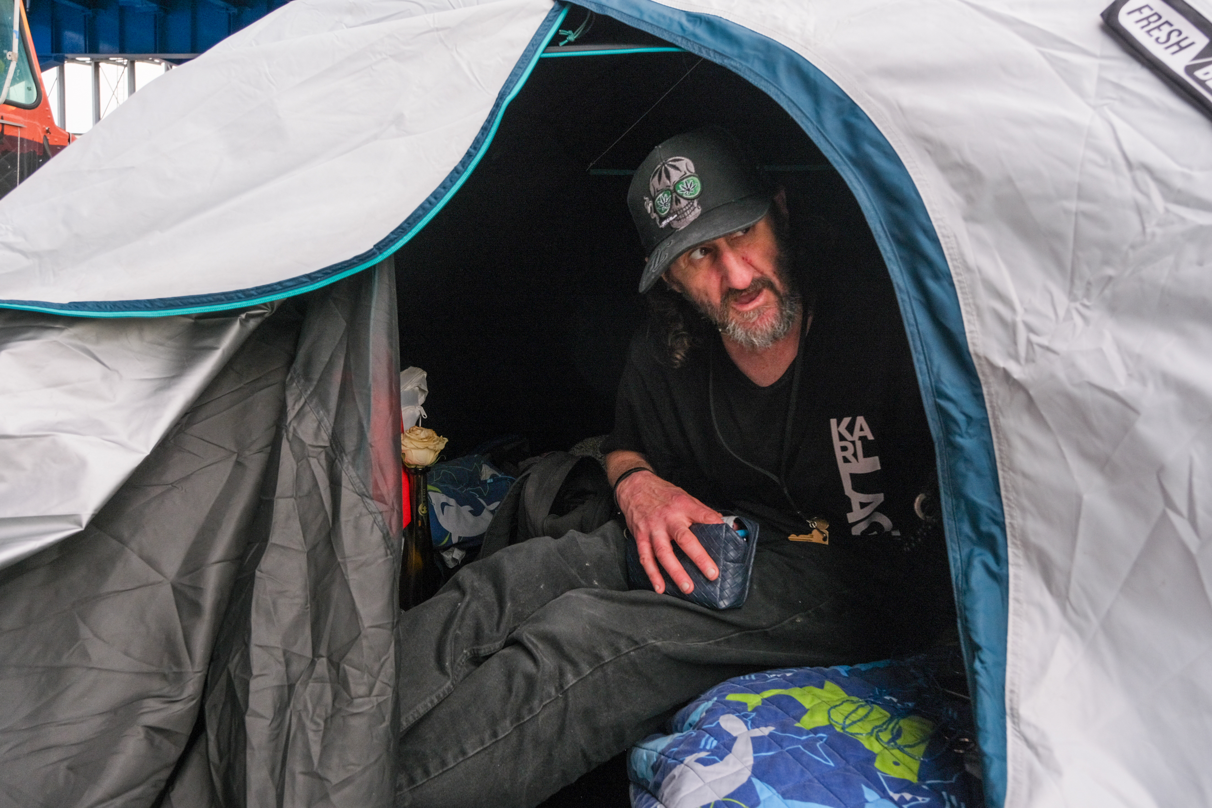 A man with a beard and cap sits inside a white and blue tent. He holds a blue wallet and rests on a colorful blanket, with a small bouquet visible beside him.