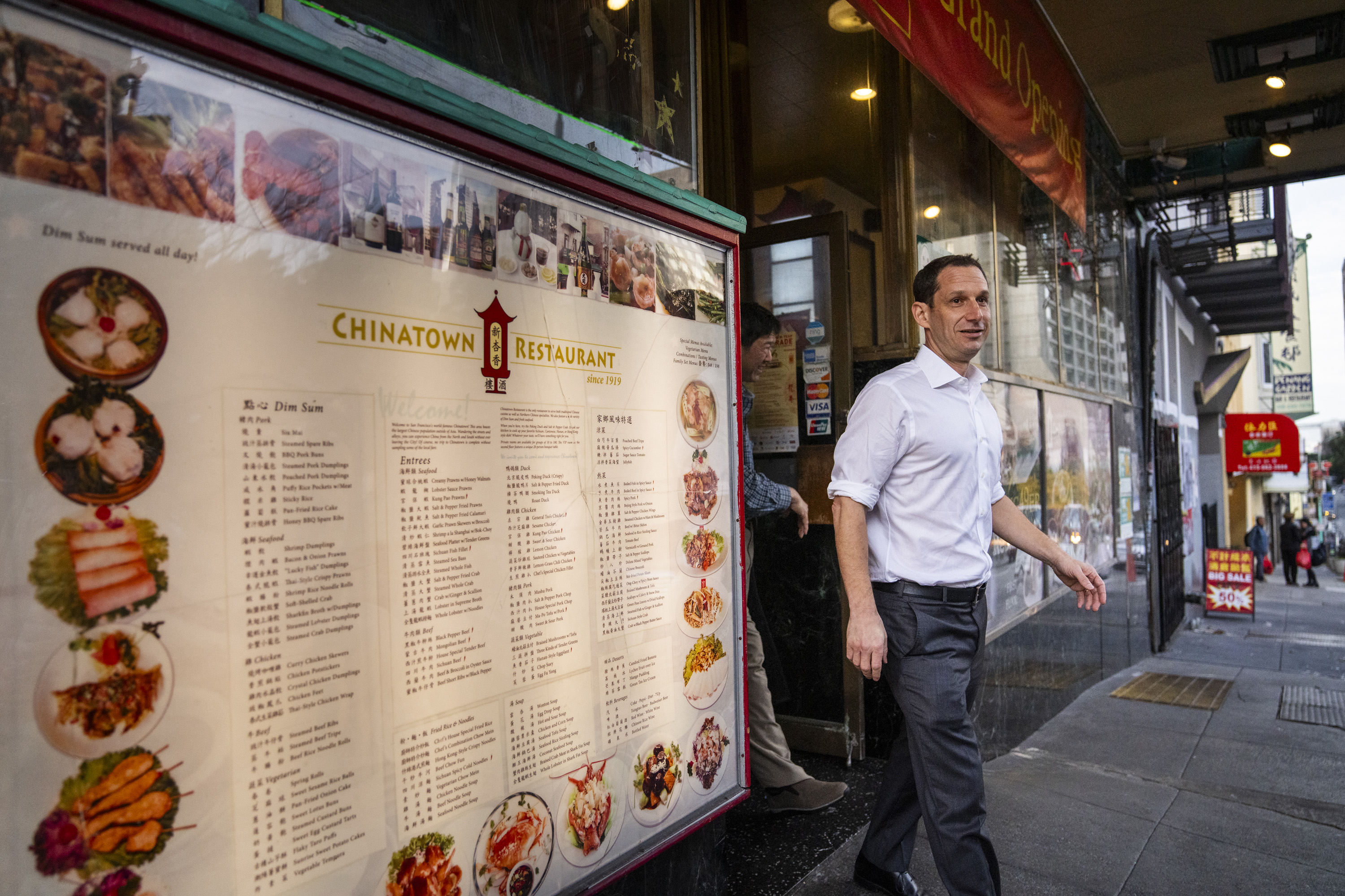 A man in a white shirt walks past a Chinatown restaurant menu displayed outside, showcasing dim sum and entrees, with colorful food images and text.