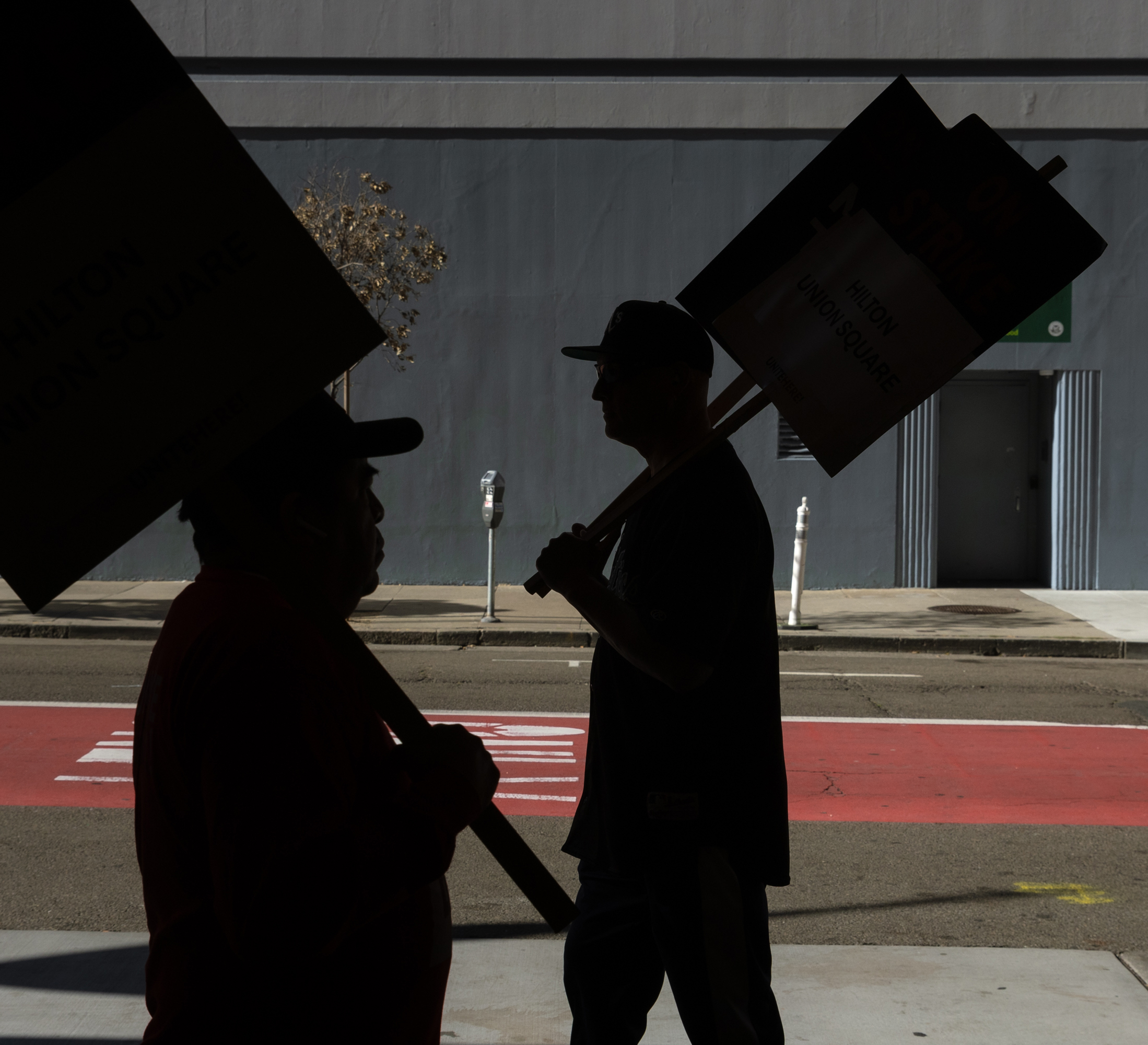 Silhouetted against a street, two people hold protest signs. The background shows a building with a grey wall, a doorway, and a sidewalk with a parking meter.