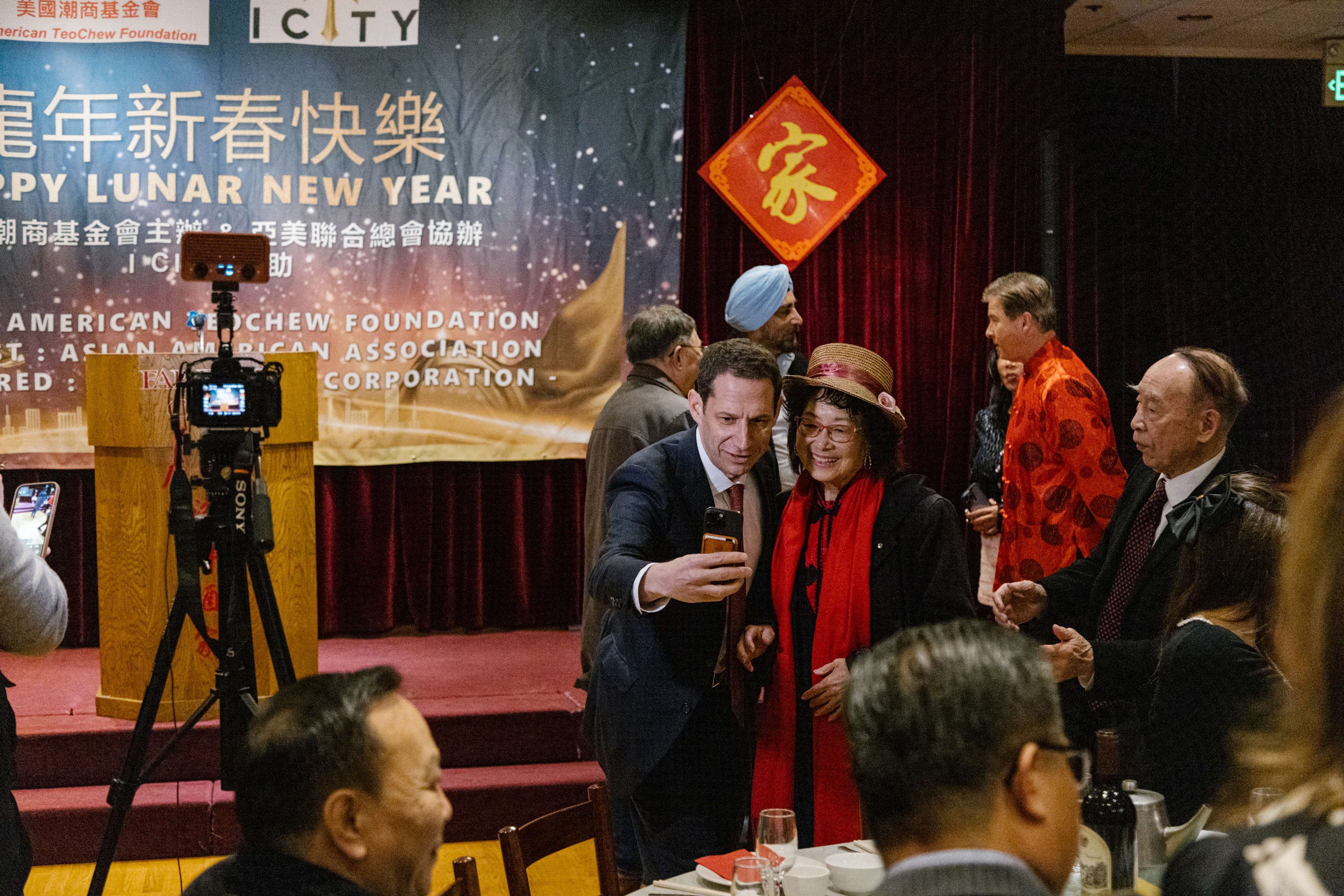 A group of people gather at a Lunar New Year event. Two people are posing for a selfie in the foreground. A festive banner and decorations are visible.