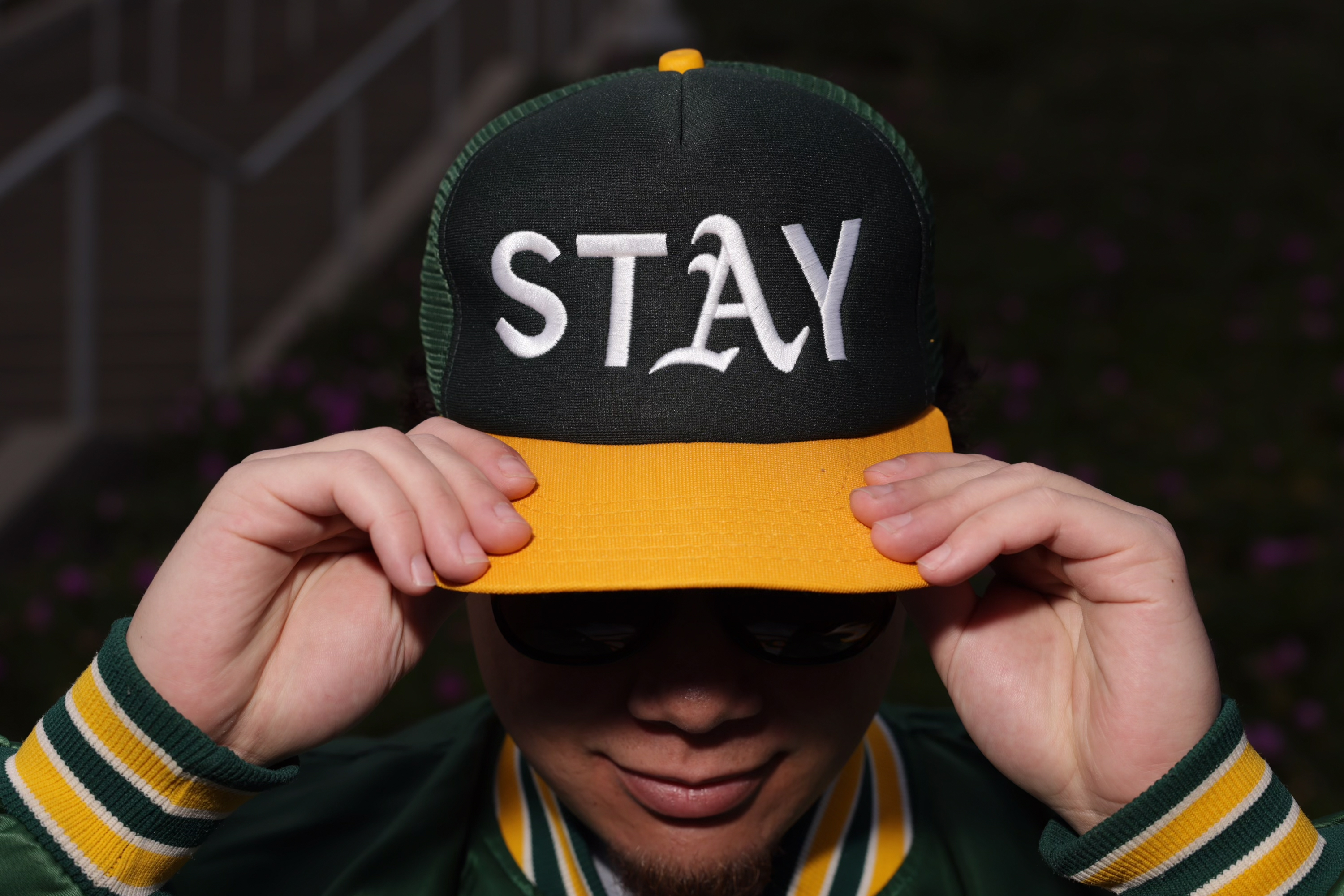 A person wearing a green and yellow cap with &quot;STAY&quot; written on it adjusts the brim. They are also wearing sunglasses and a green striped jacket.