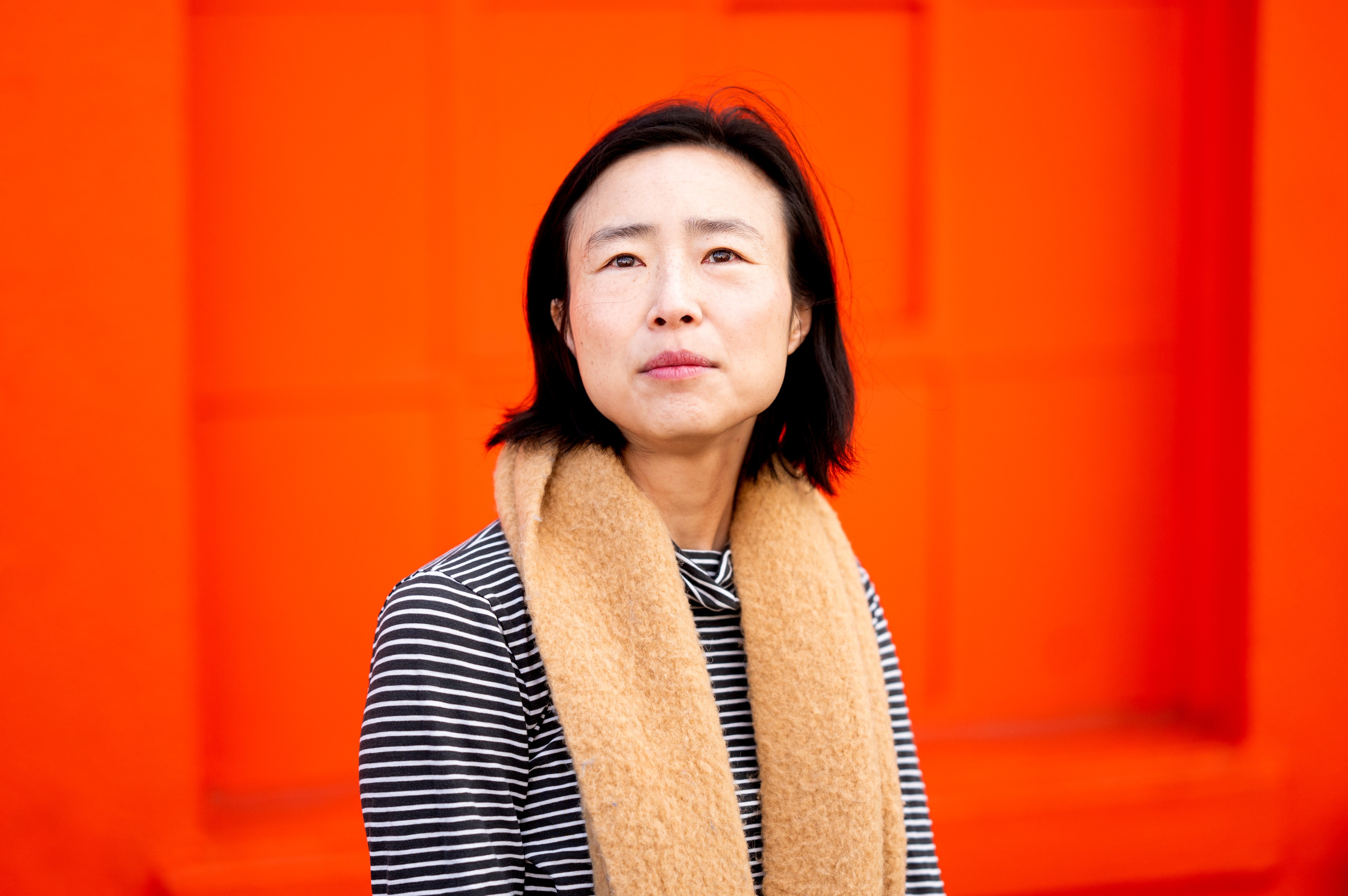 A person with short black hair wears a striped shirt and a tan scarf, standing in front of a bright orange background, looking slightly upward.