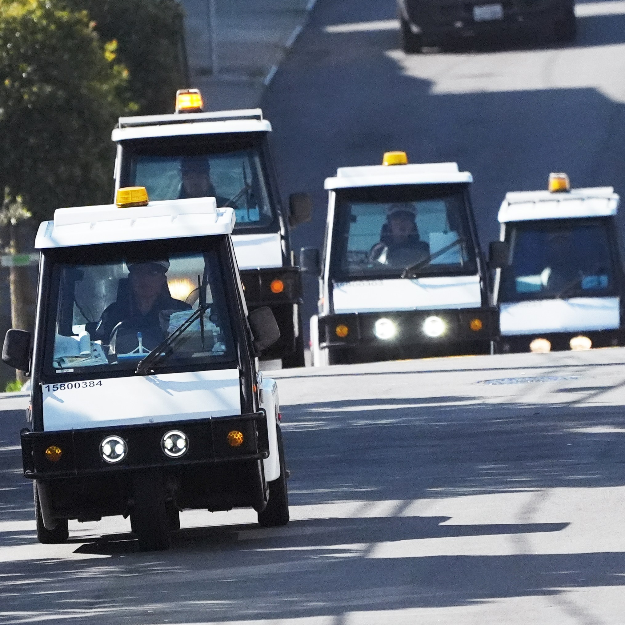 A convoy of four small, white utility vehicles with yellow lights on top drives down a sunny, tree-lined street. Shadows and parked cars are visible.