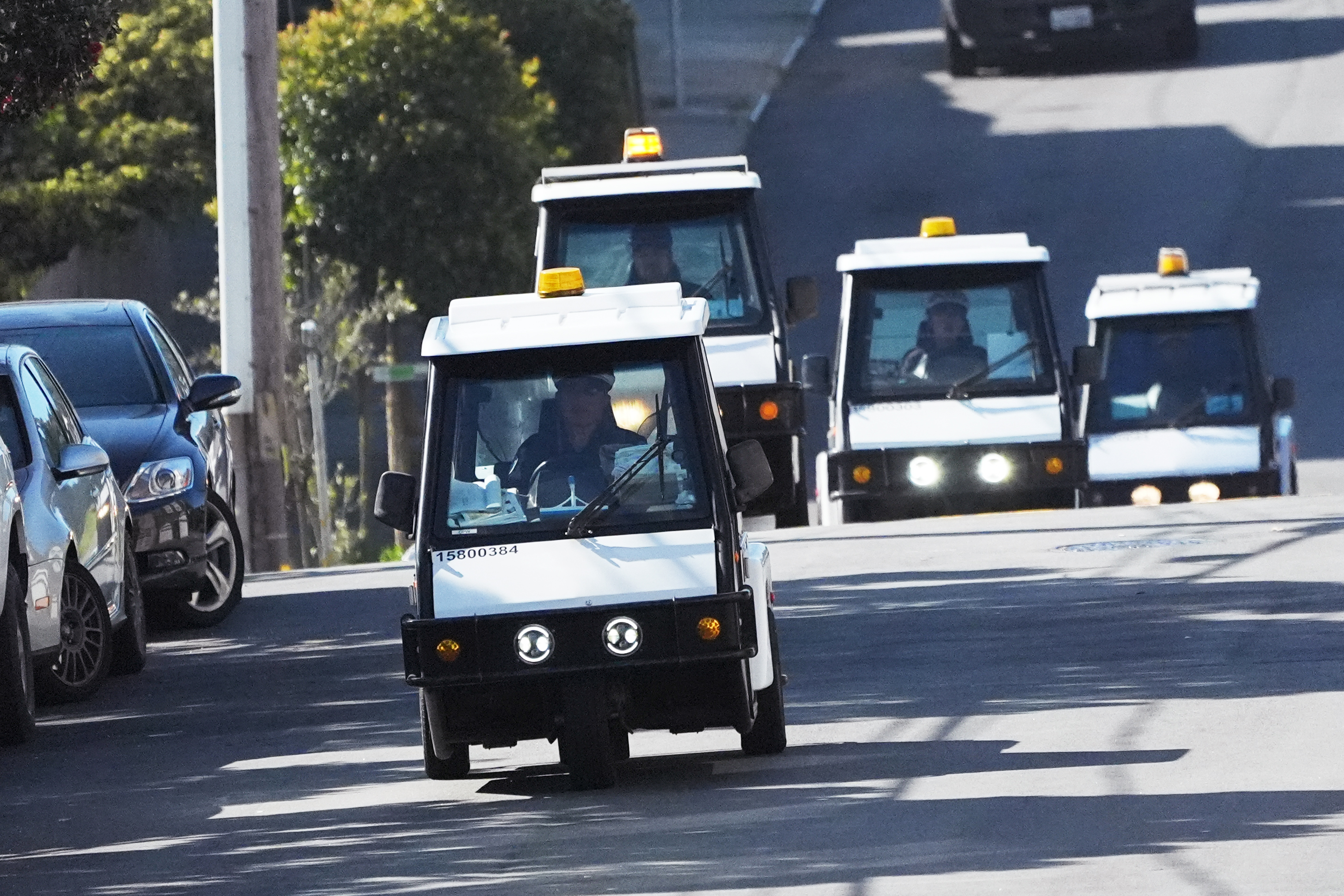 A convoy of four small, white utility vehicles with yellow lights on top drives down a sunny, tree-lined street. Shadows and parked cars are visible.