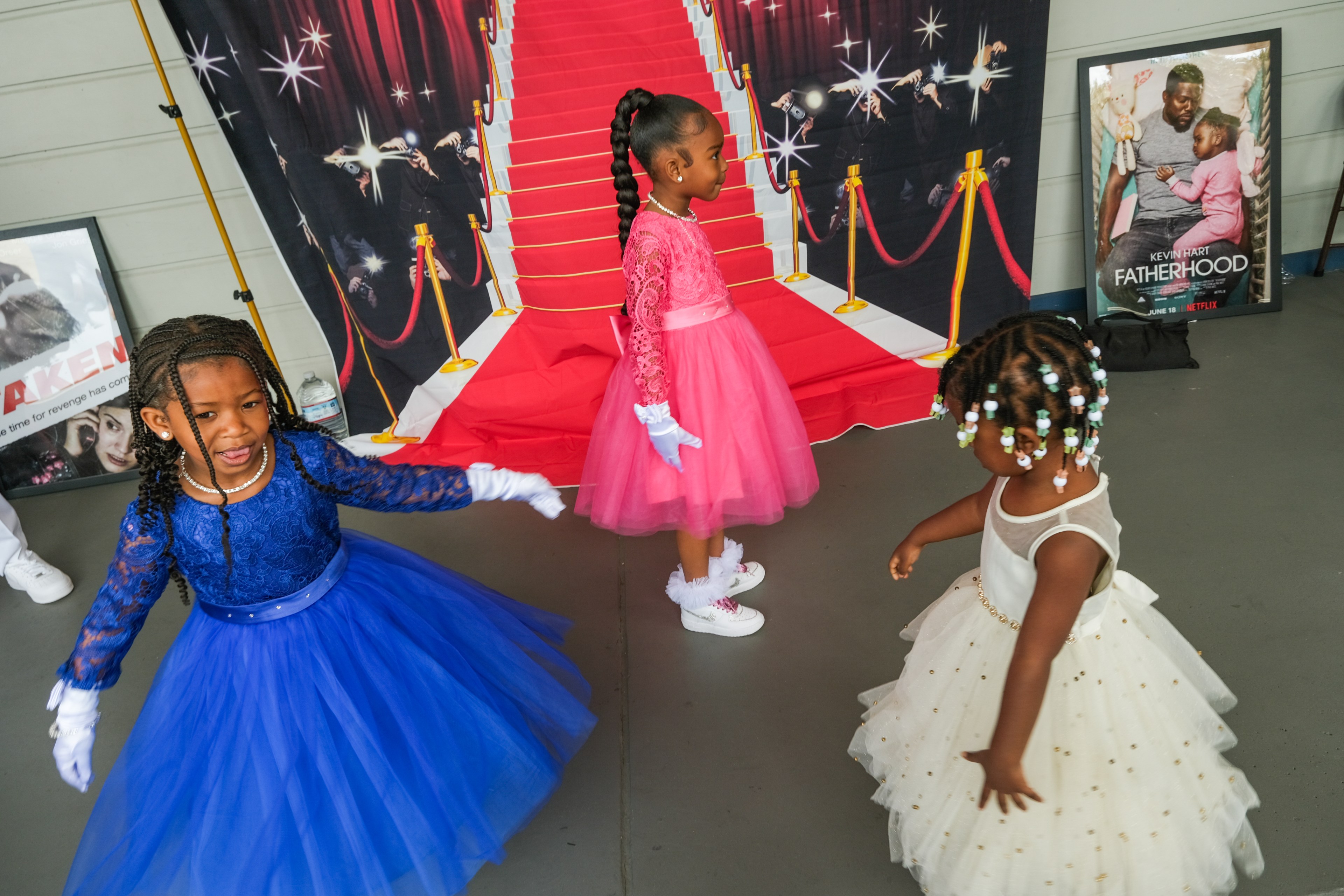 Three little girls in fancy dresses—blue, pink, and white—playfully dance on a red-carpet-themed backdrop, with movie posters displayed behind them.