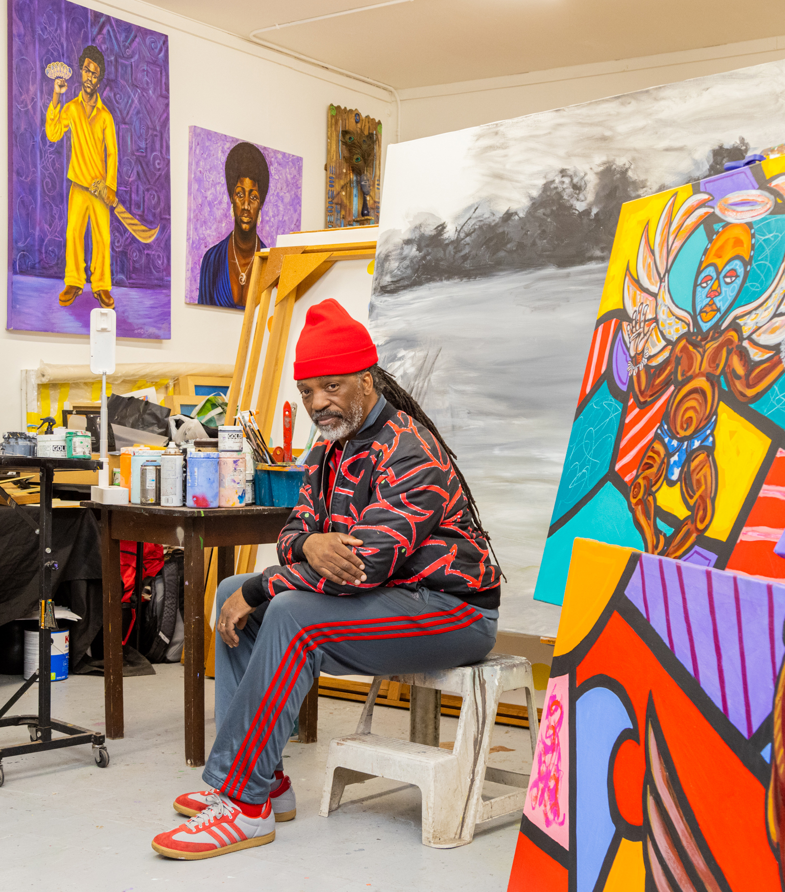 A person wearing a red beanie and sportswear sits in an art studio. The room is filled with vibrant abstract and portrait paintings and a table with paint cans.