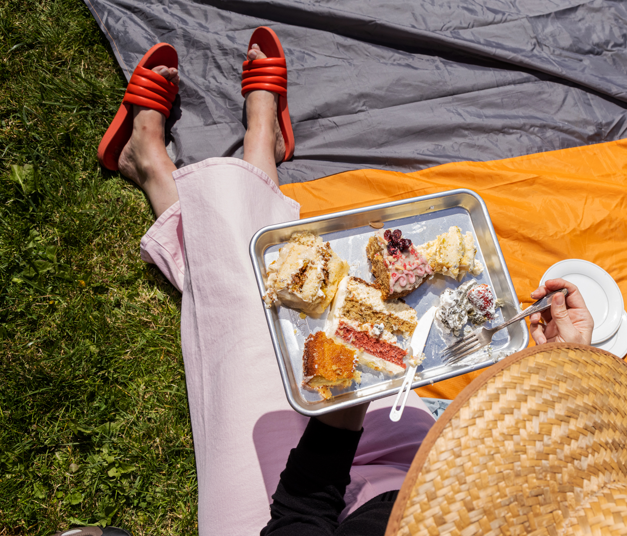 A person with pink pants and red sandals is sitting on grass, holding a tray of assorted cake slices. They're also holding a straw hat and a fork.