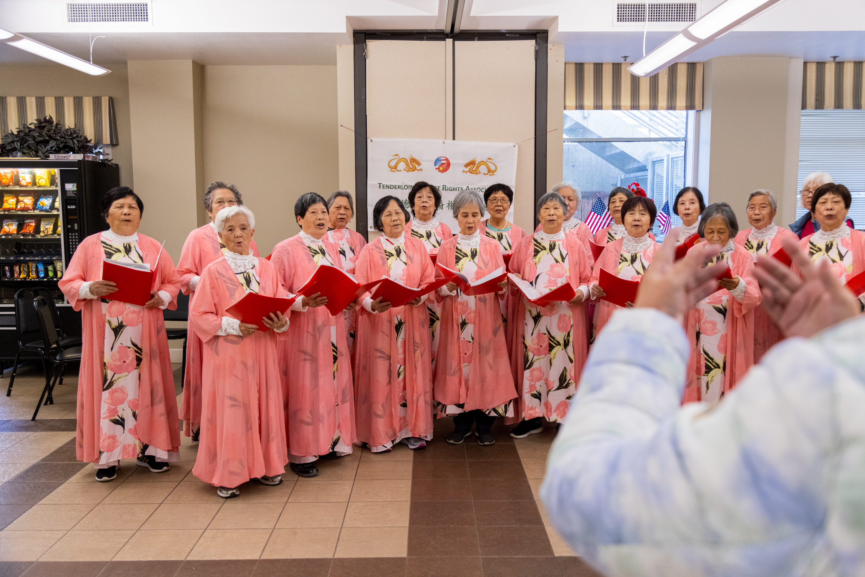 A choir of elderly women in pink floral dresses holds red folders while singing. They are in a room with striped curtains and a vending machine. An instructor gestures.