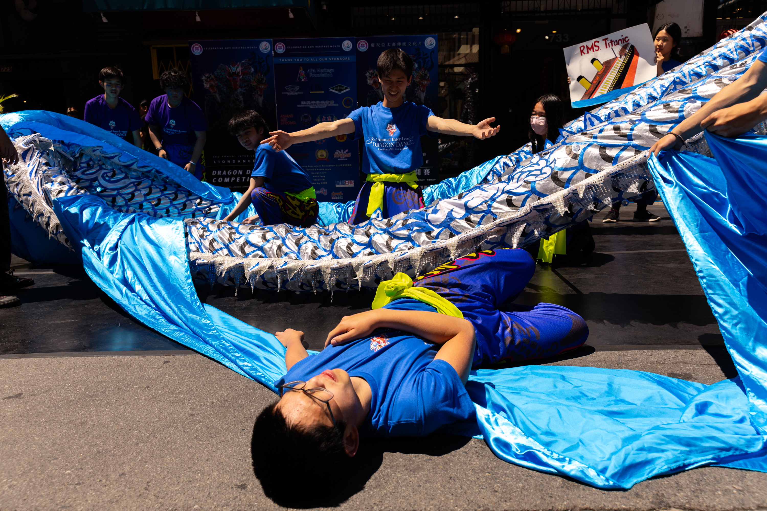 A group of people in blue costumes hold up an intricate, flowing fabric dragon. One person lies beneath it, while others smile and gesture energetically.