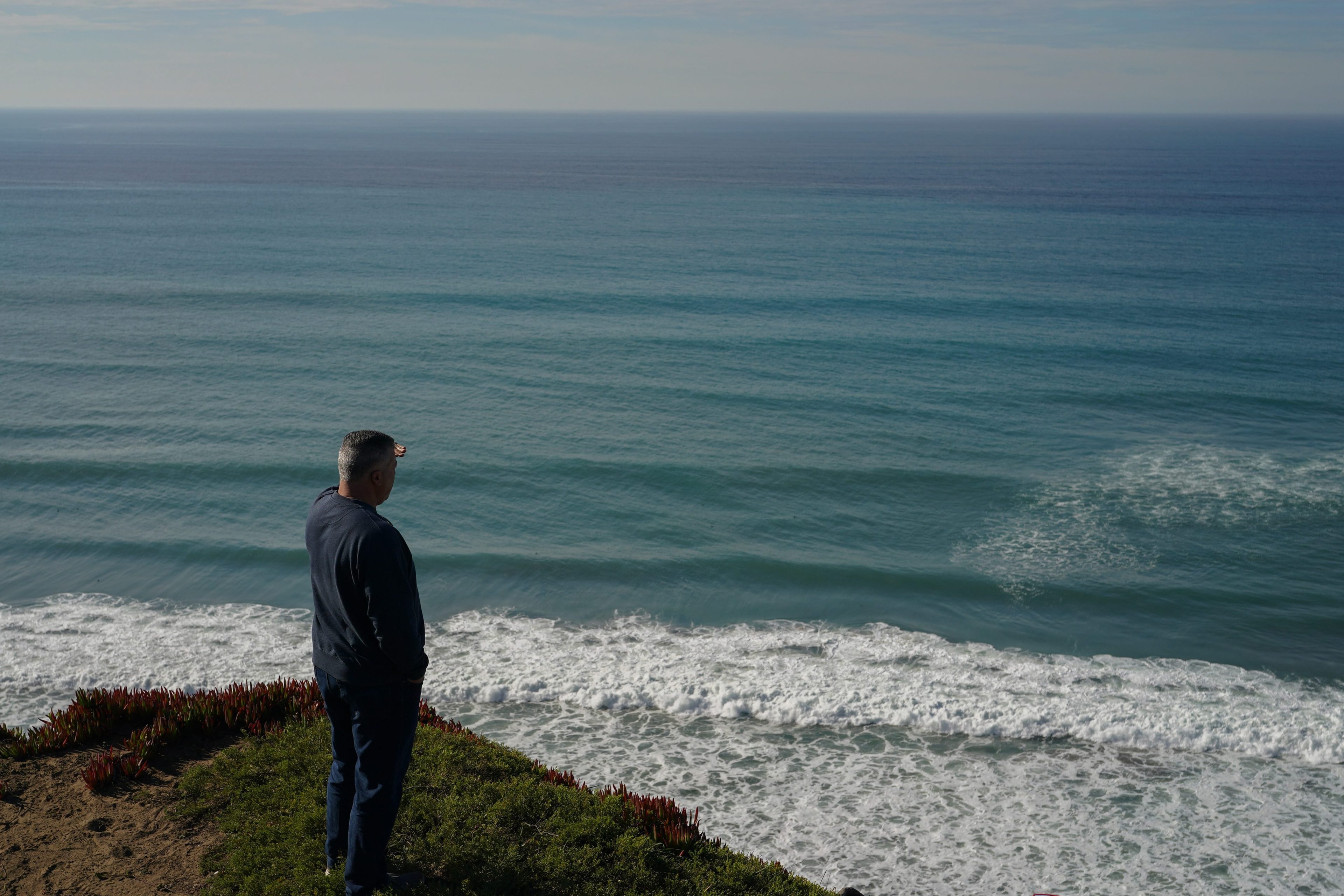 A person stands on a grassy cliff, gazing at the vast ocean. The waves gently crash onto the shore below under a clear blue sky.