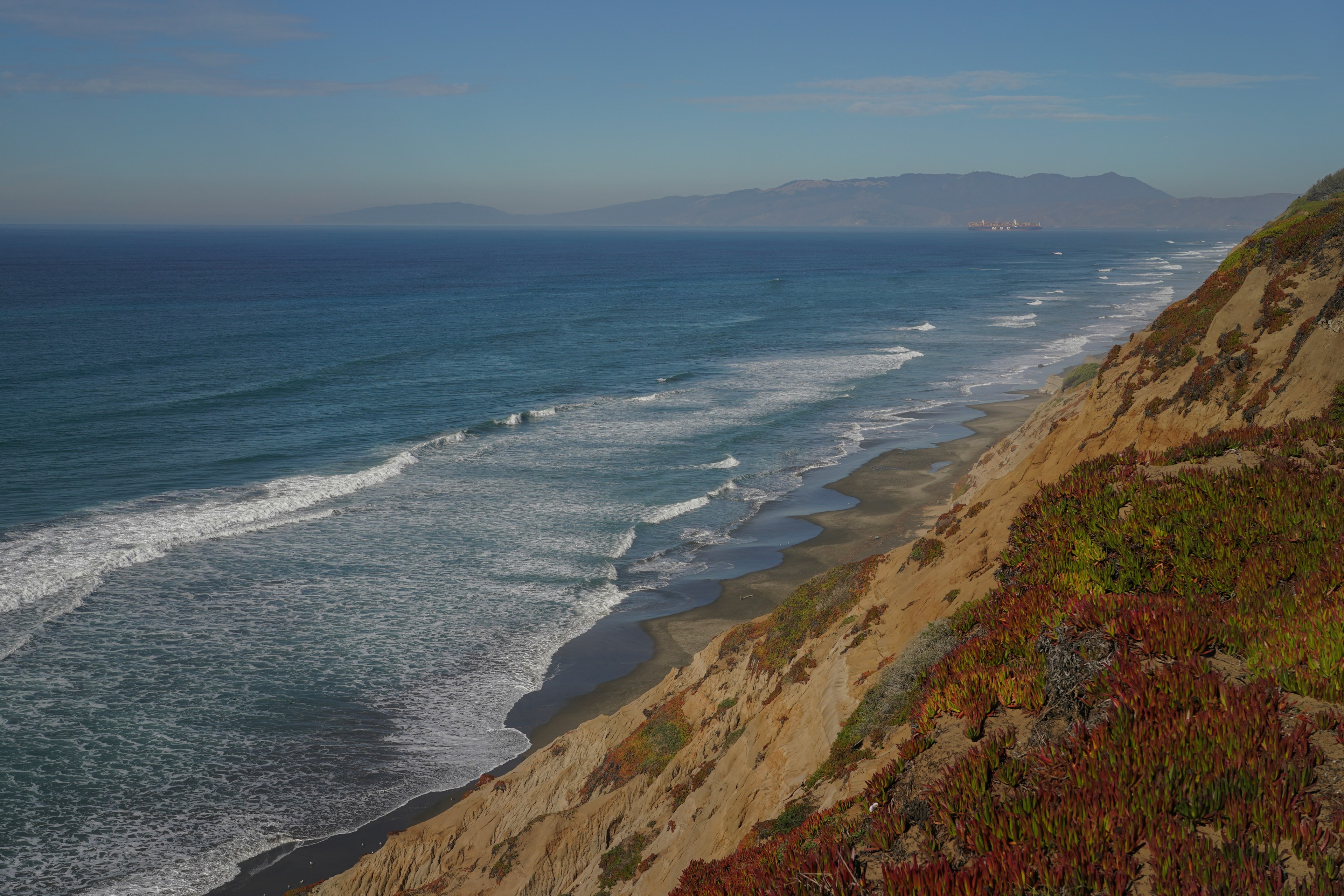 A coastline view with waves gently crashing onto a sandy beach bordered by steep cliffs covered in greenery, set against a distant mountain backdrop.