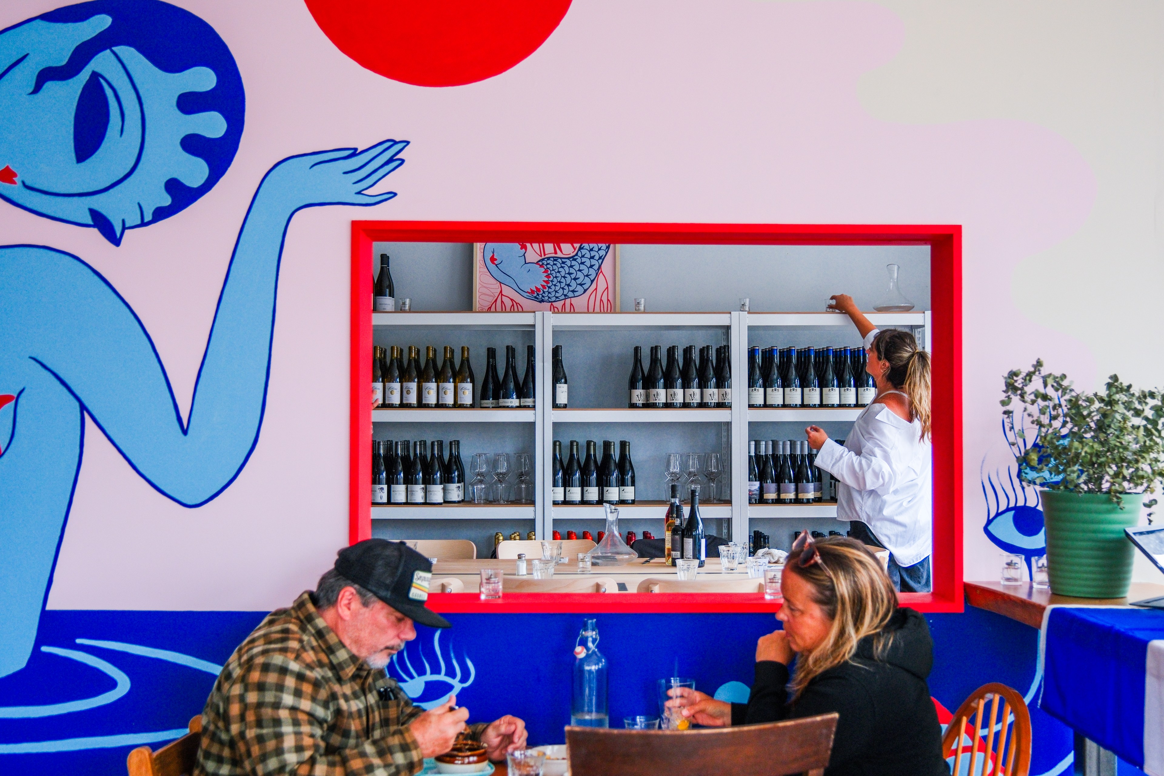 A vibrant café with bold wall art; a woman reaches for a bottle on shelves filled with wine behind a red-framed opening, while two people dine in the foreground.