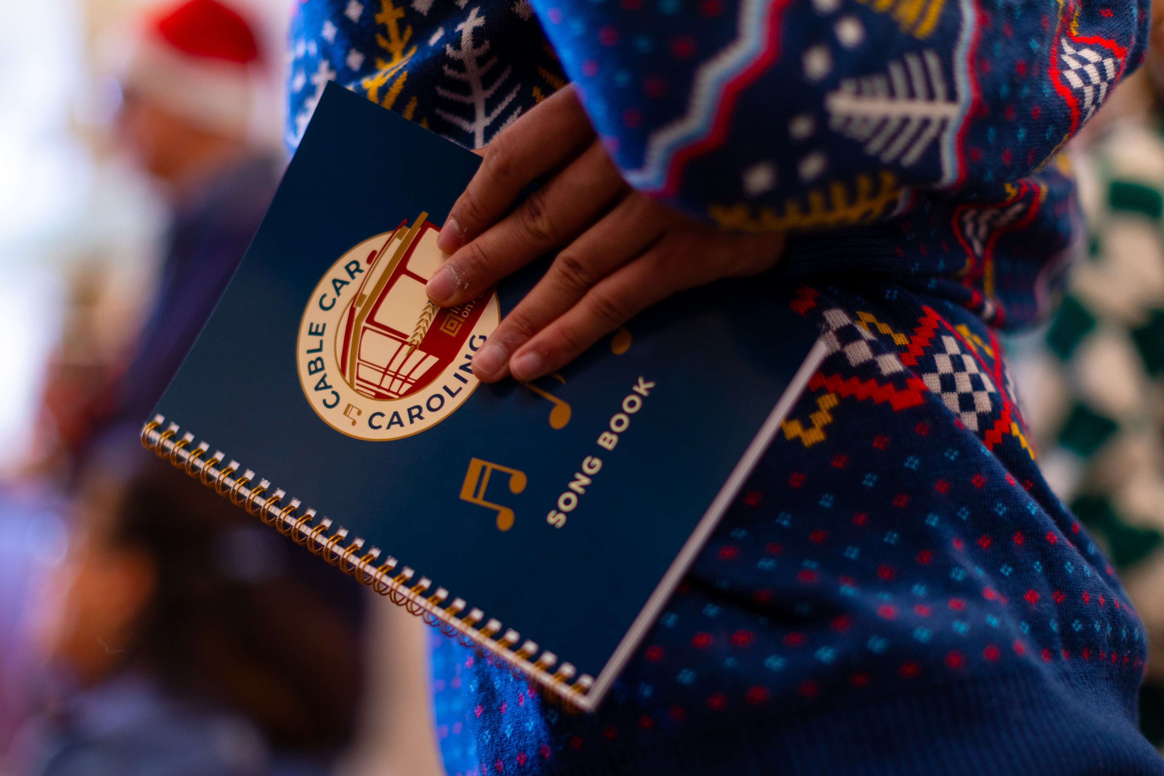 A person wearing a colorful sweater holds a blue songbook with a cable car logo. In the background, someone in a Santa hat is blurred.