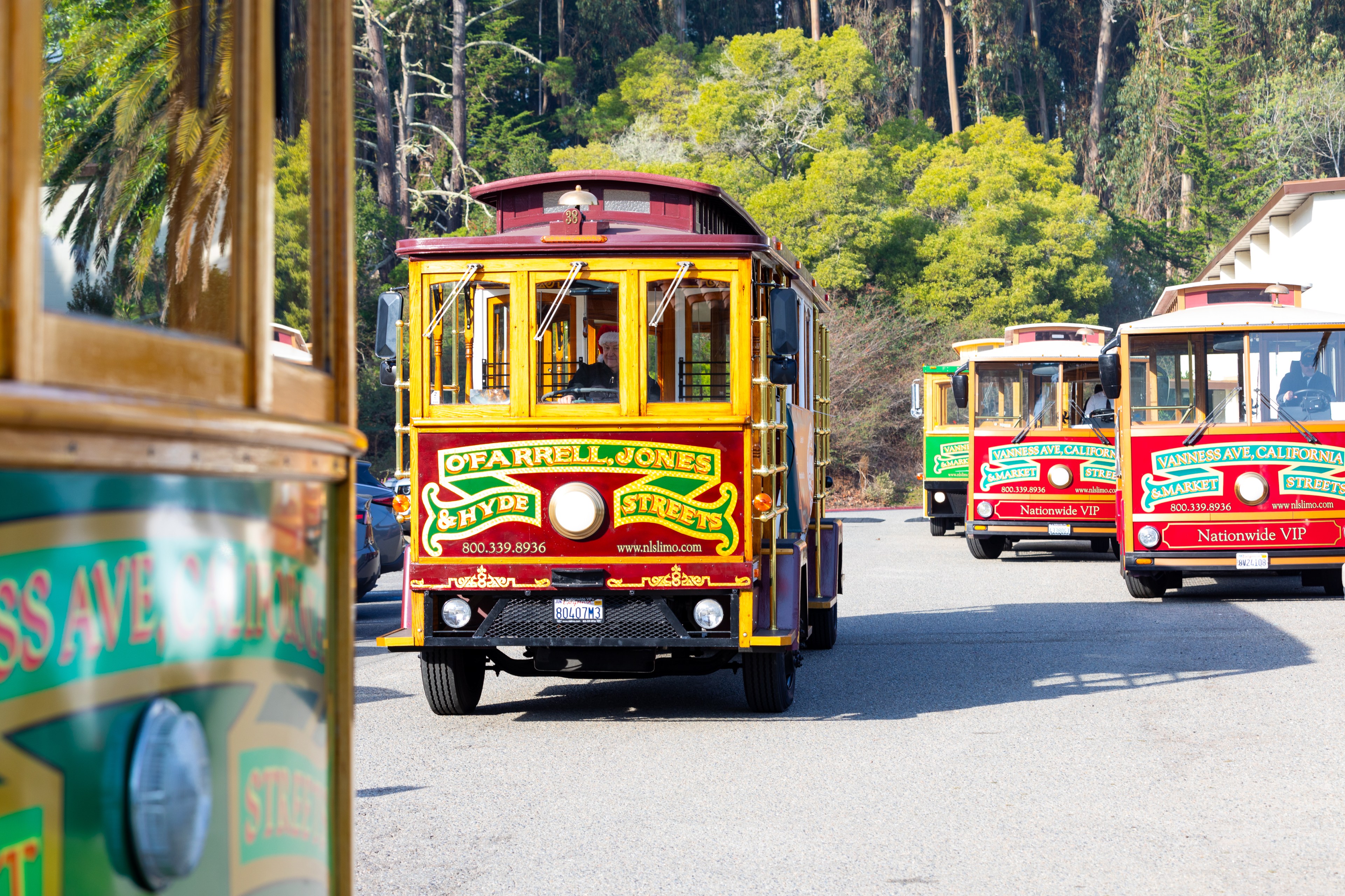 The image shows several colorful trolley buses on a road lined with trees. The central trolley is red and yellow with &quot;O'Farrell, Jones &amp; Hyde&quot; written on the front.