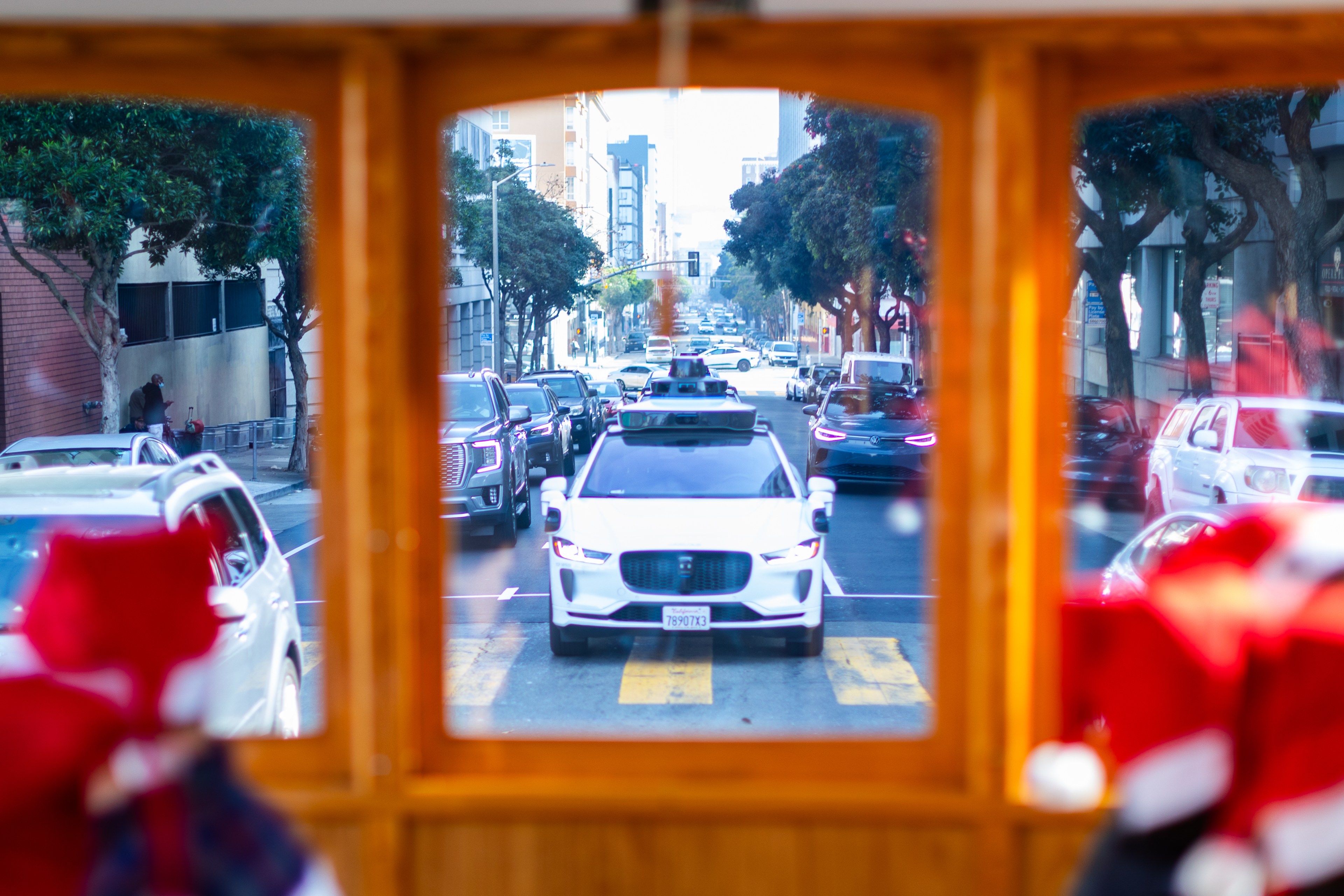 The image shows a busy urban street with a self-driving white car leading a line of vehicles, framed by the interior of a streetcar. Trees and buildings line the road.