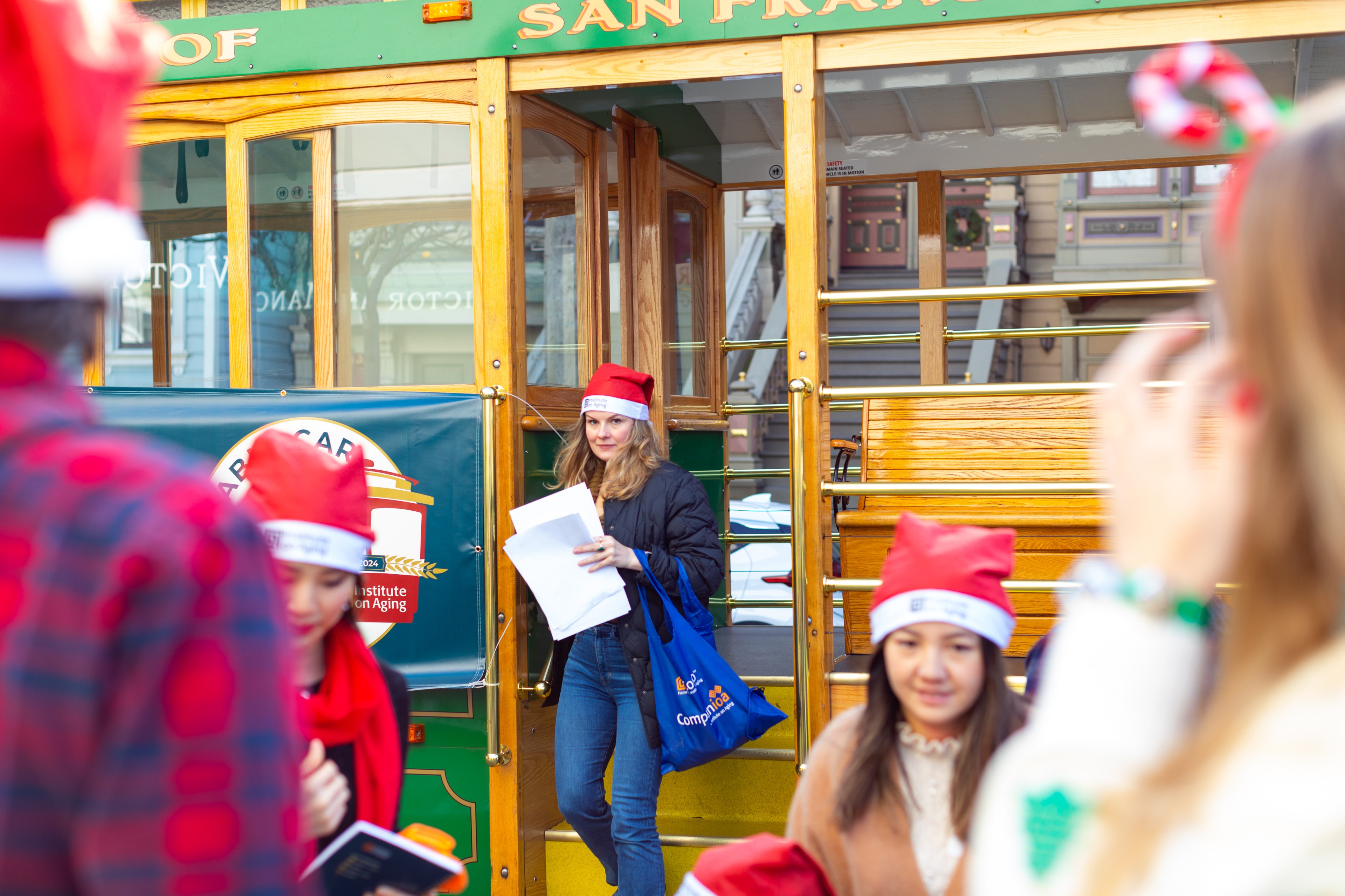 A group of people in red Santa hats are gathered around a classic San Francisco cable car. One person holds papers, and there's a festive atmosphere.