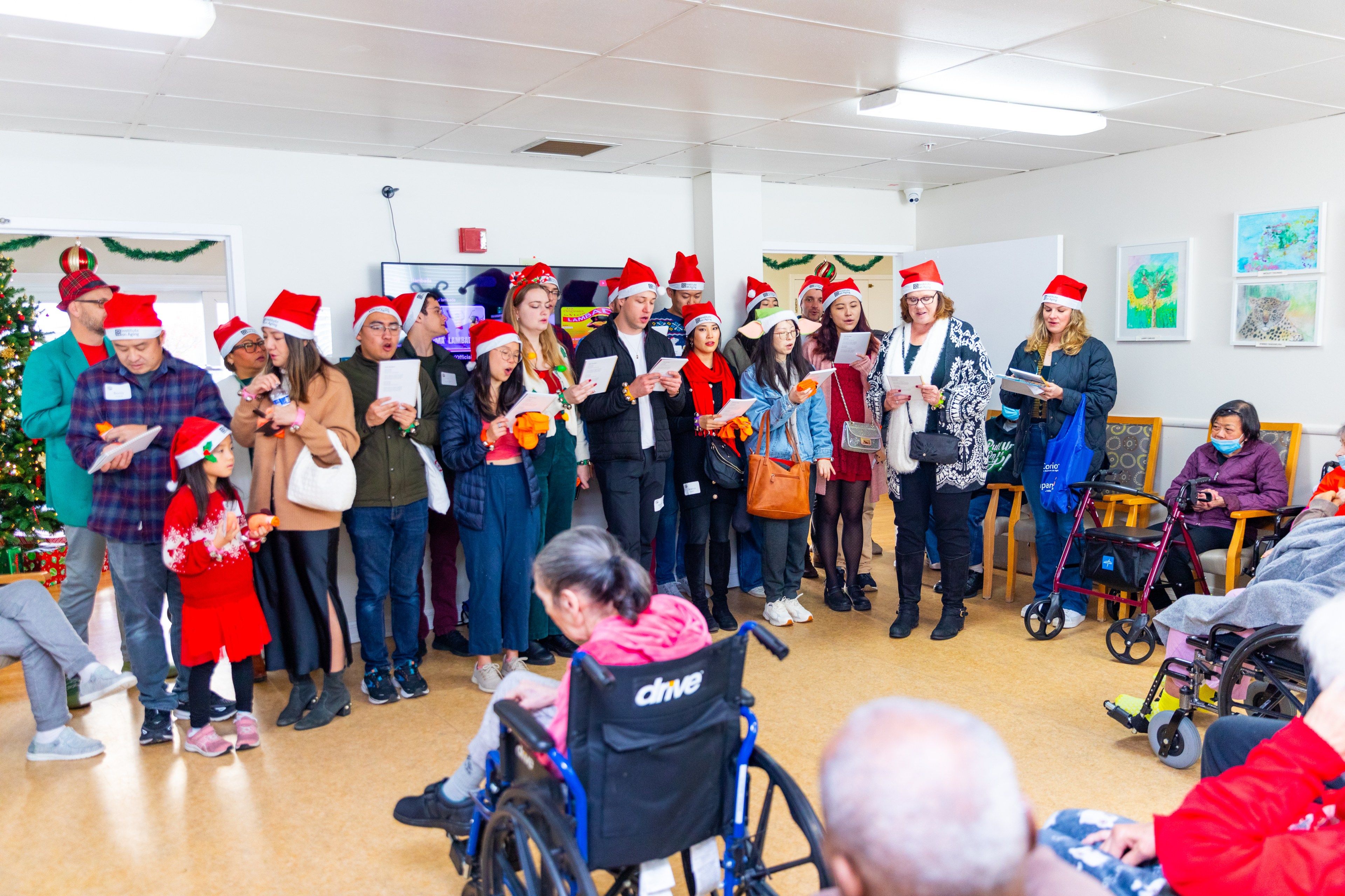 A group of people wearing Santa hats sings together in a festive indoor setting, while several elderly individuals, some in wheelchairs, watch and listen.