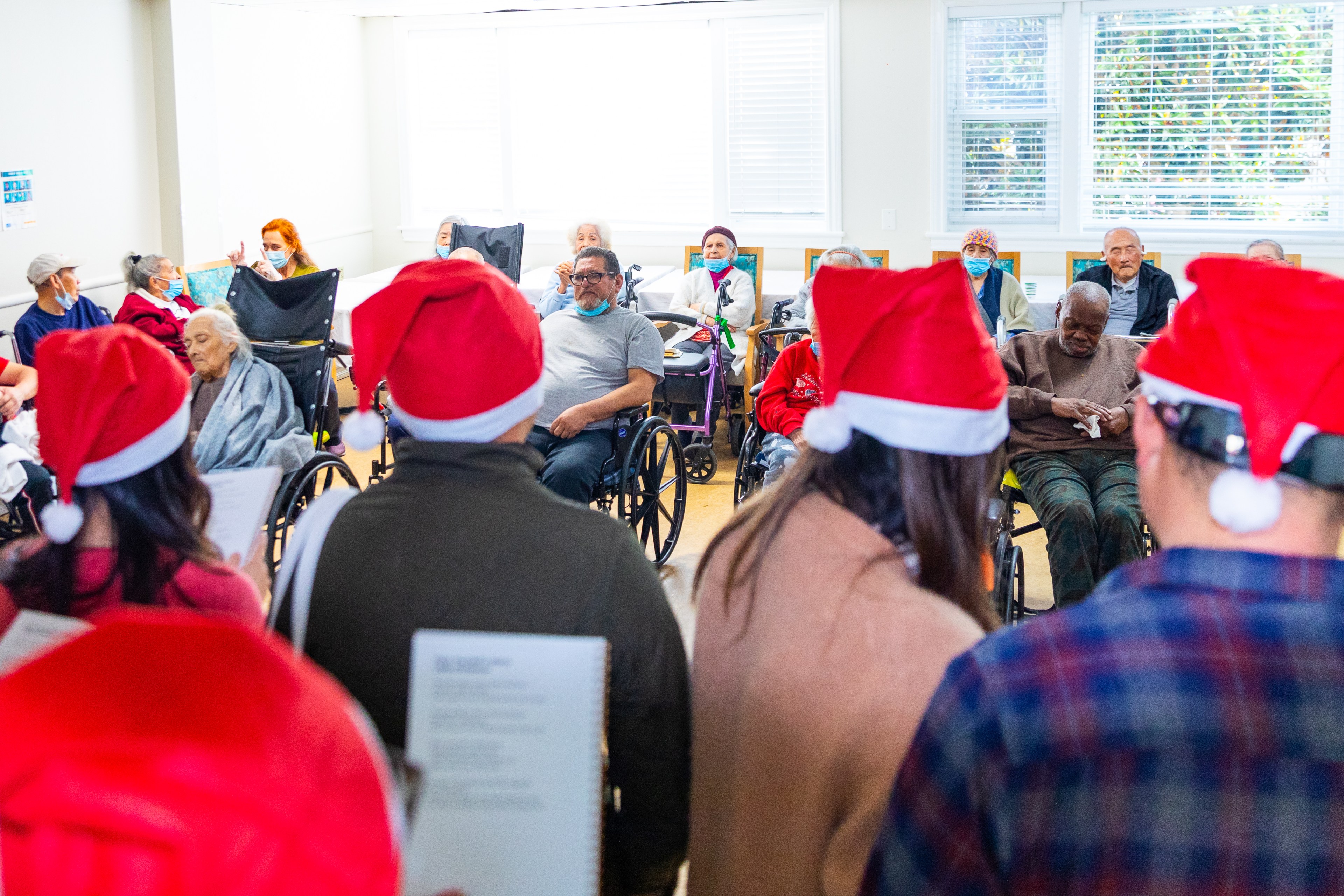 People wearing Santa hats are singing to an audience of elderly individuals in wheelchairs, some wearing masks, in a brightly lit room.